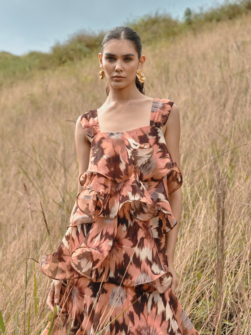 Woman in a ruffled brown and pink patterned dress standing in tall grass.