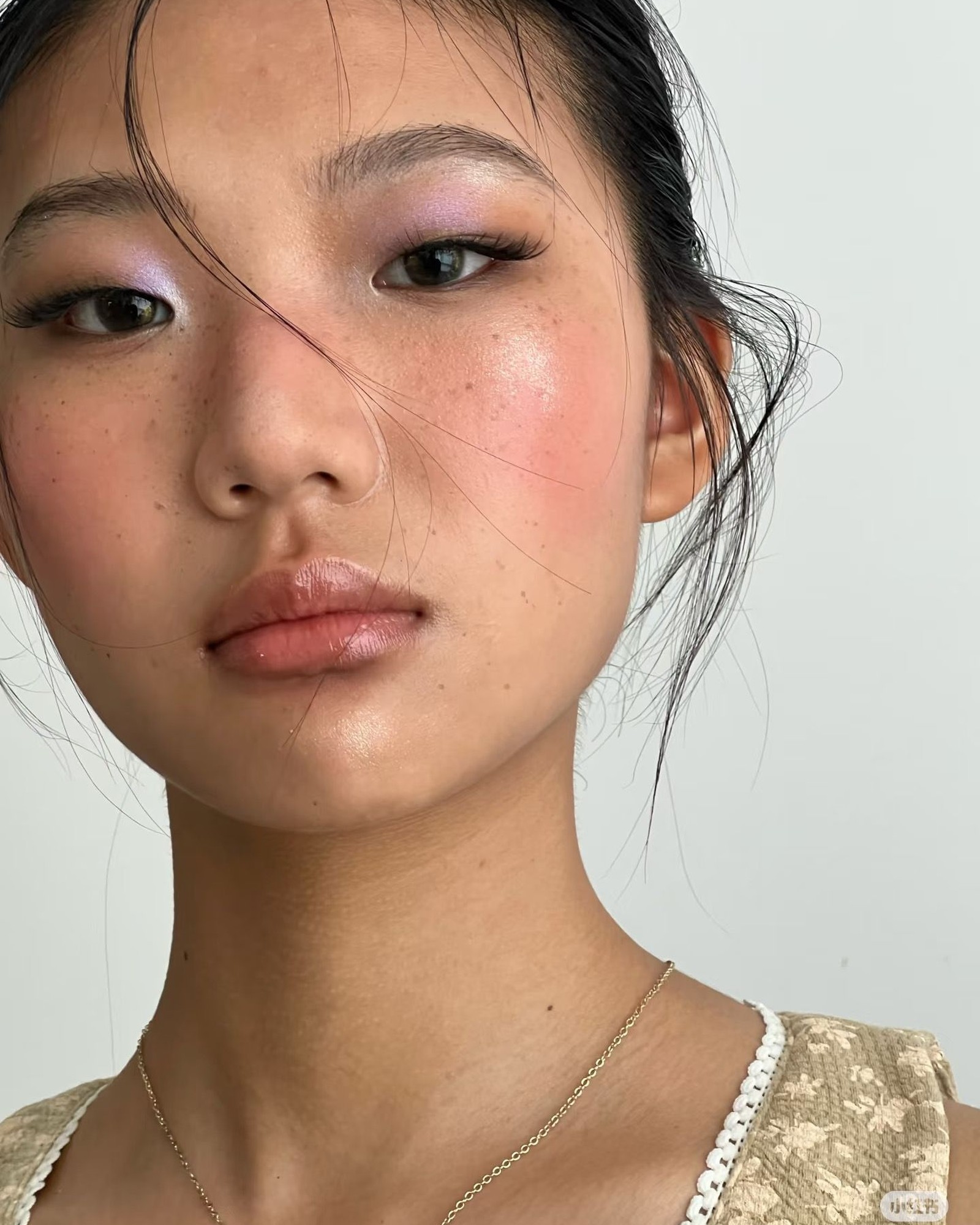Close portrait of a freckled woman with lilac eyeshadow, glossy pink cheeks and lips, wet-look hair and a thin gold chain