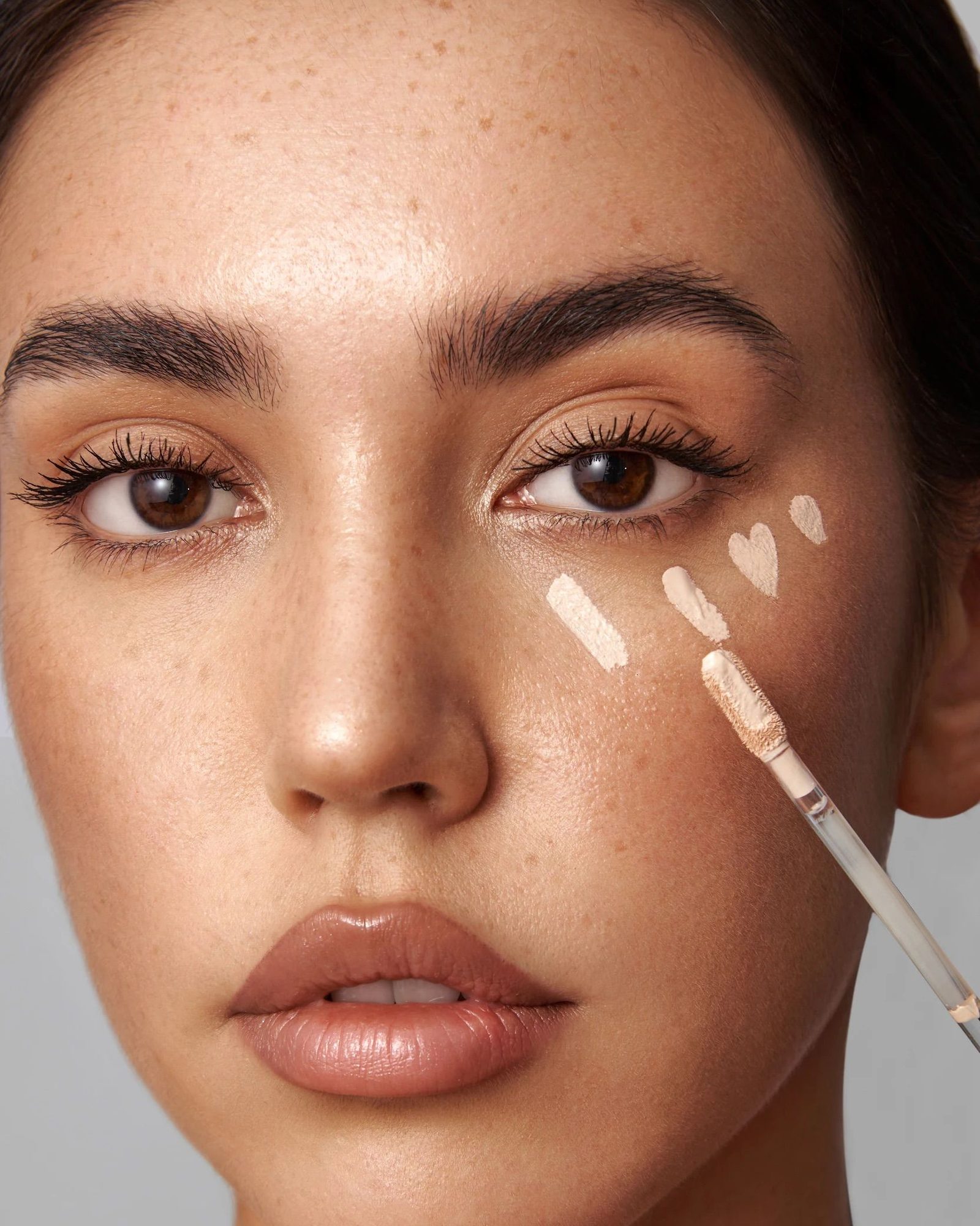 Close-up of a freckled woman with swipes of concealer on her cheek and a doe-foot applicator wand