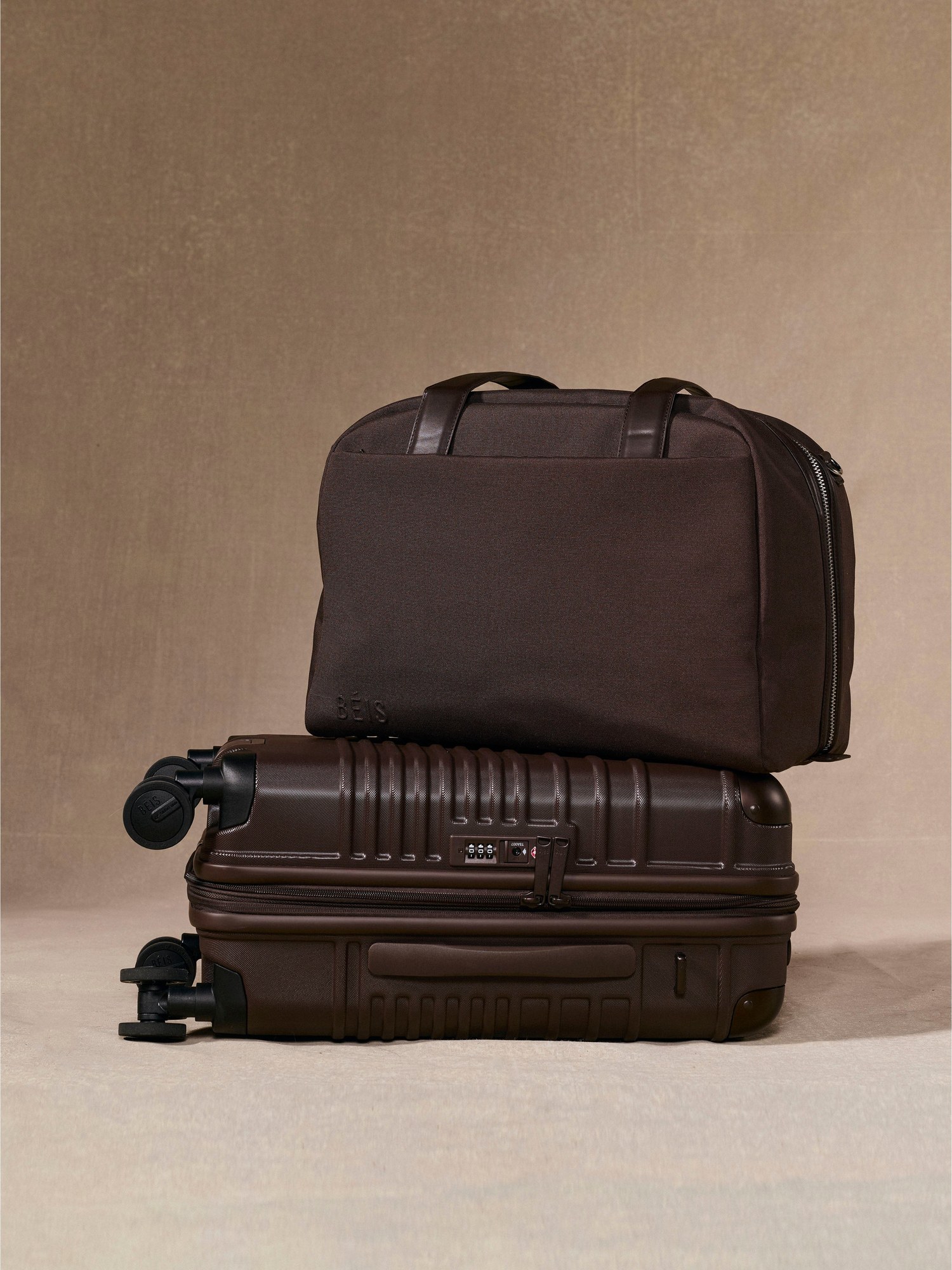 Dark brown Beis duffle stacked on a carry-on against a warm beige wall