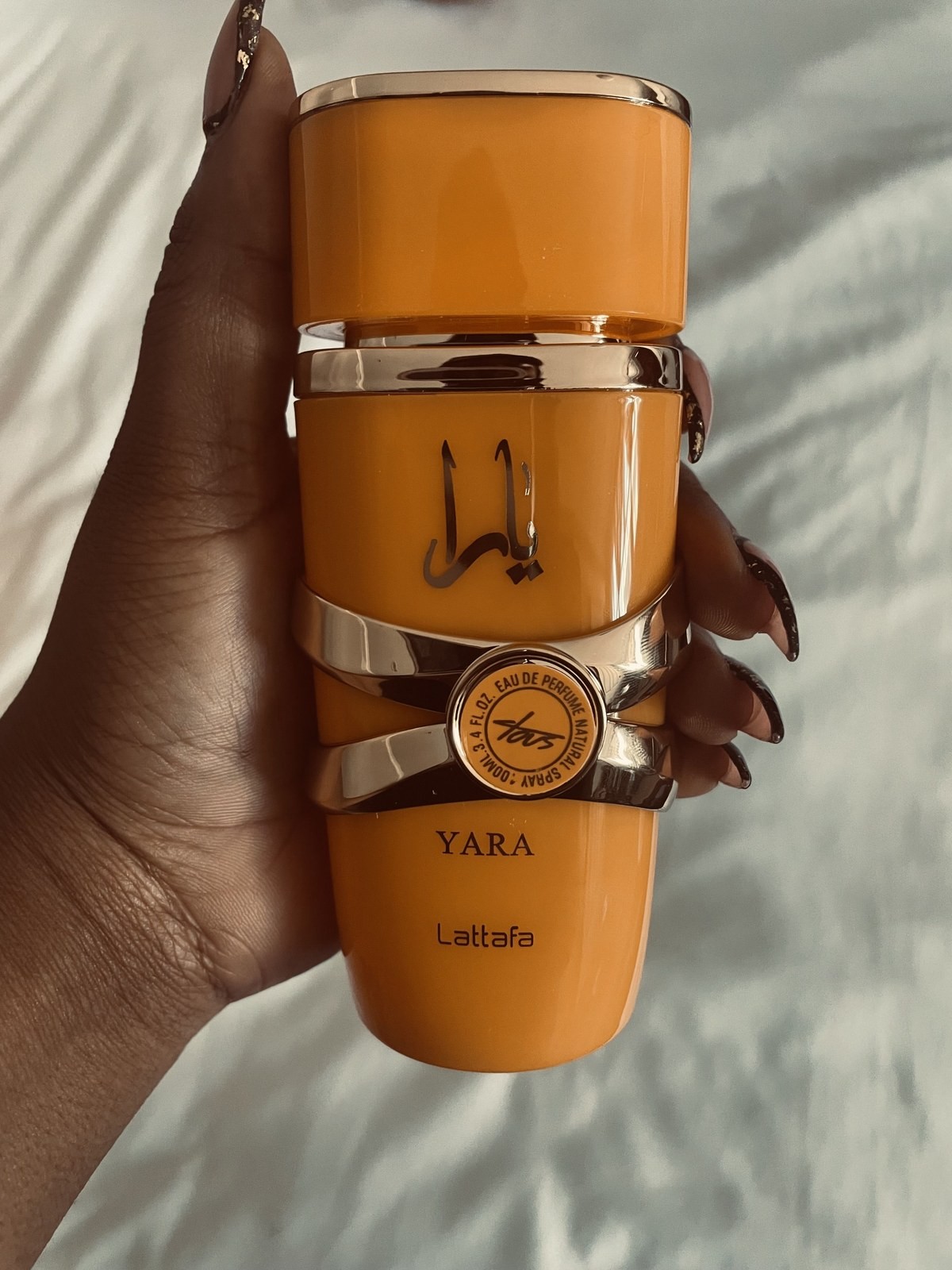 Hand holding an orange Lattafa Yara Tous perfume bottle against a white bedspread