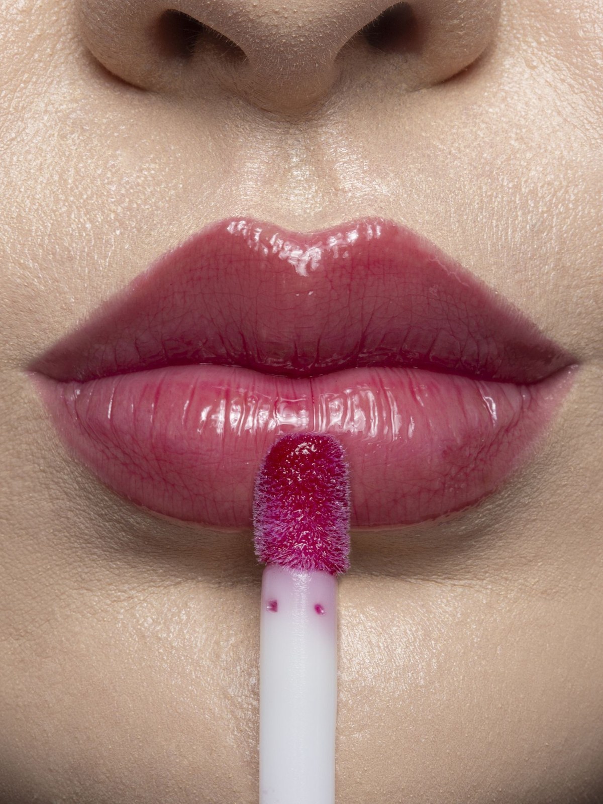 Close up of glossy berry lips with a magenta lip oil applicator held just below the mouth