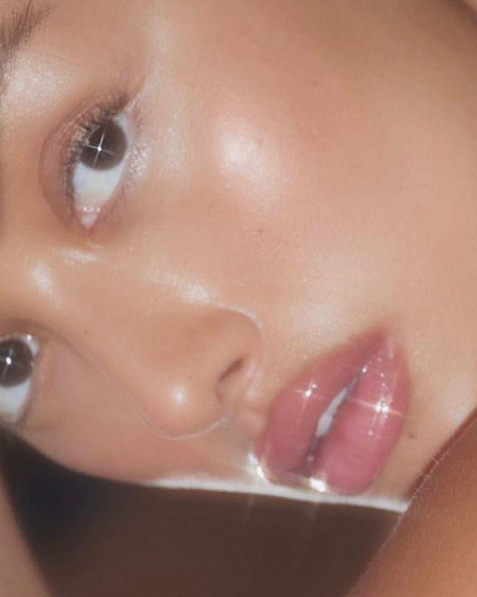 Extreme close-up of a face tilted on its side with a dewy highlight on the cheekbone and glossy pink lips with sparkle highlights