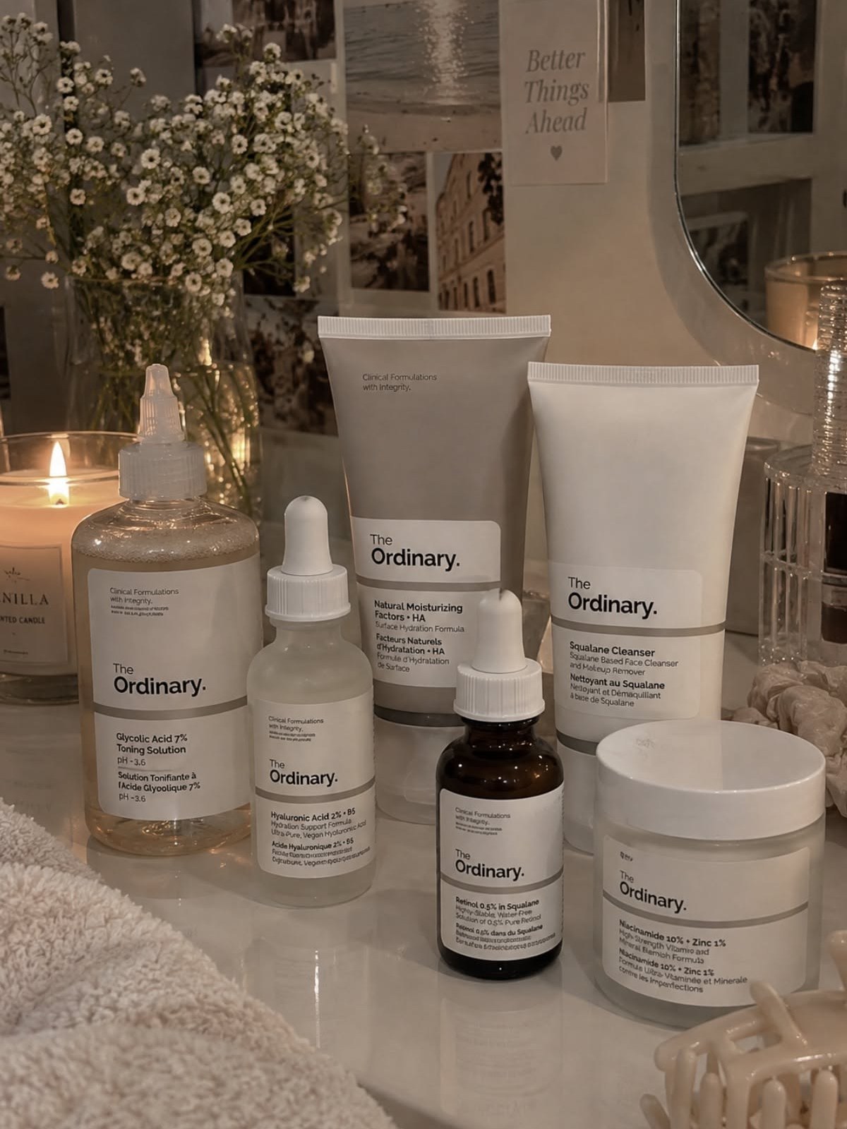 A bathroom counter by candlelight with The Ordinary Retinol 0.5% in Squalane, Natural Moisturizing Factors + HA, Squalane Cleanser, Glycolic Acid 7% and Niacinamide 10% + Zinc 1%, beside a vase of baby's breath.
