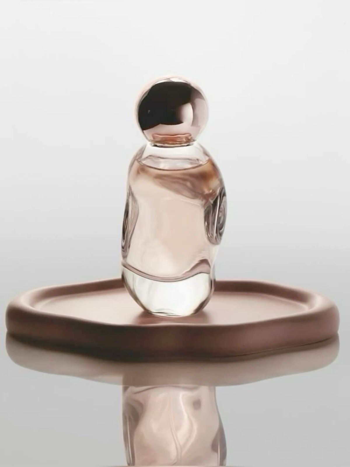 Pink-tinted glass perfume bottle with a rose gold ball cap on a sculpted rose-brown dish