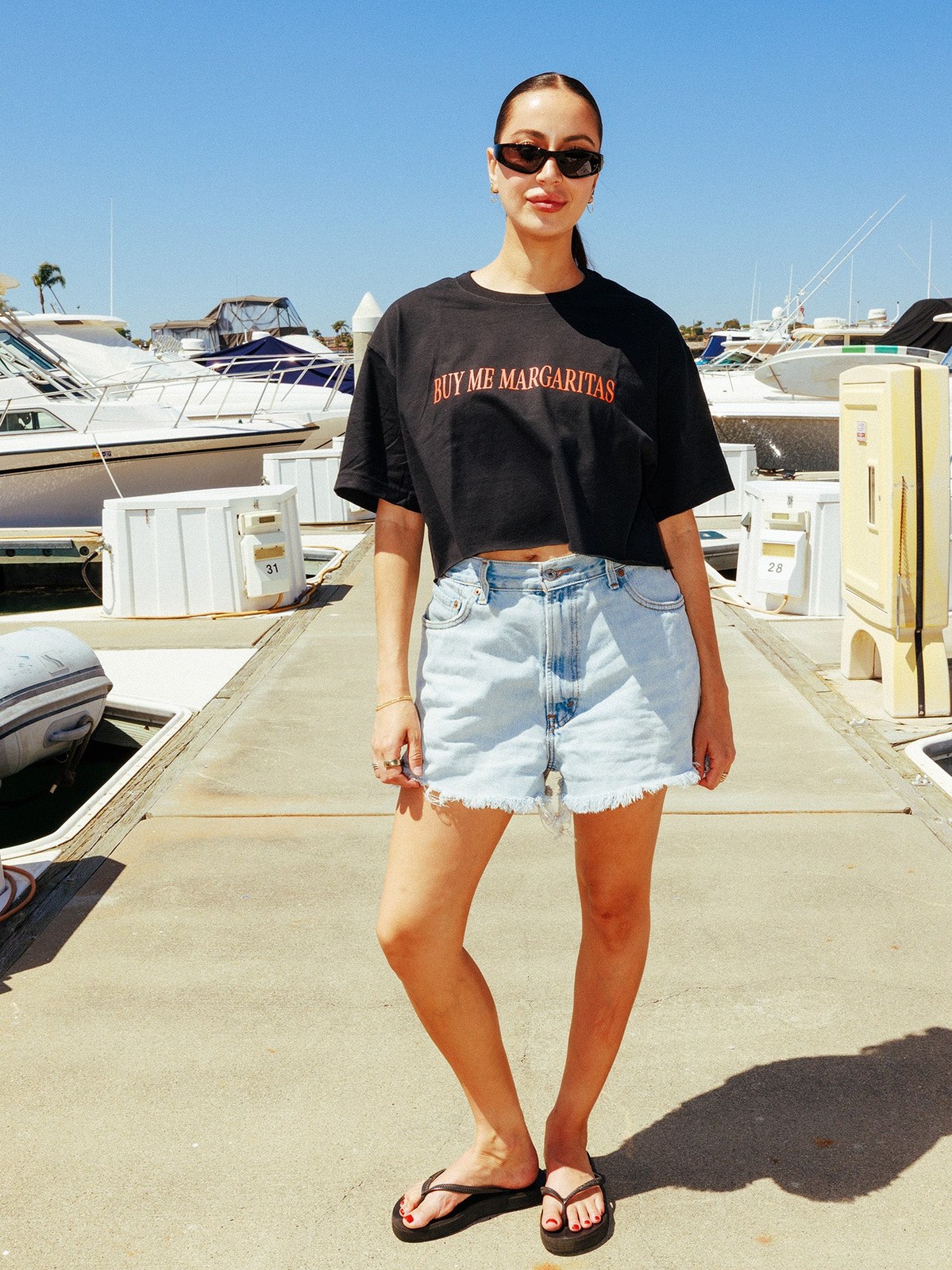 Woman in a black Motel Margarita crop tee and denim shorts at a marina