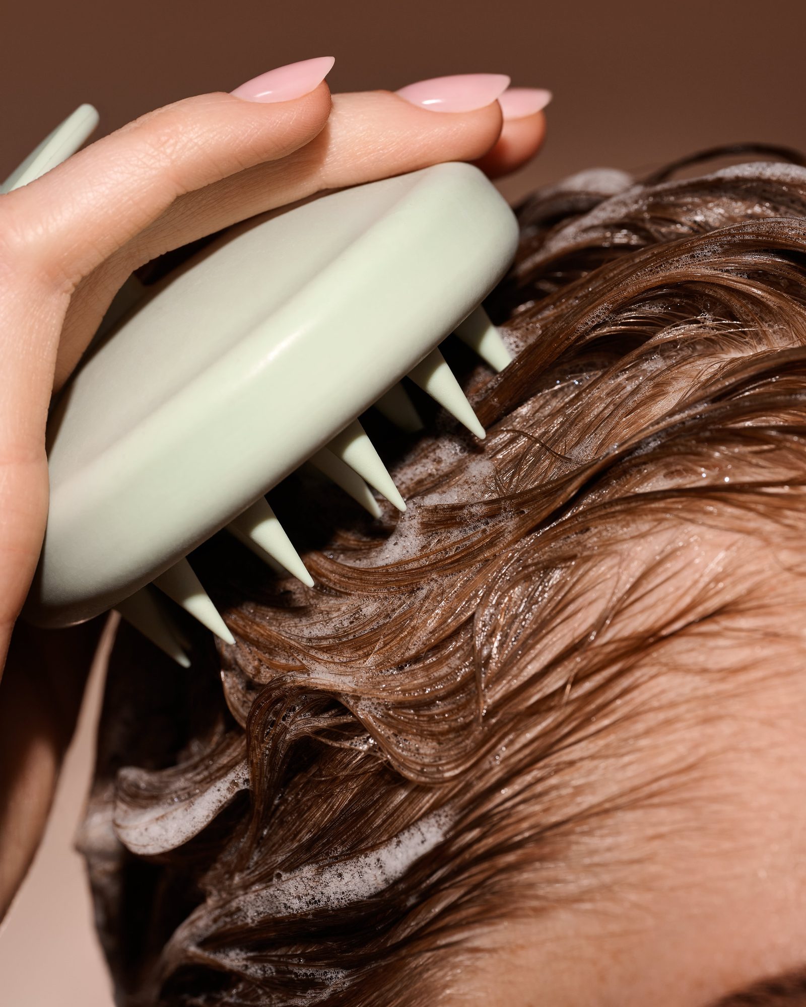 Close-up of a hand using a pale green silicone scalp massager on soapy wet brown hair