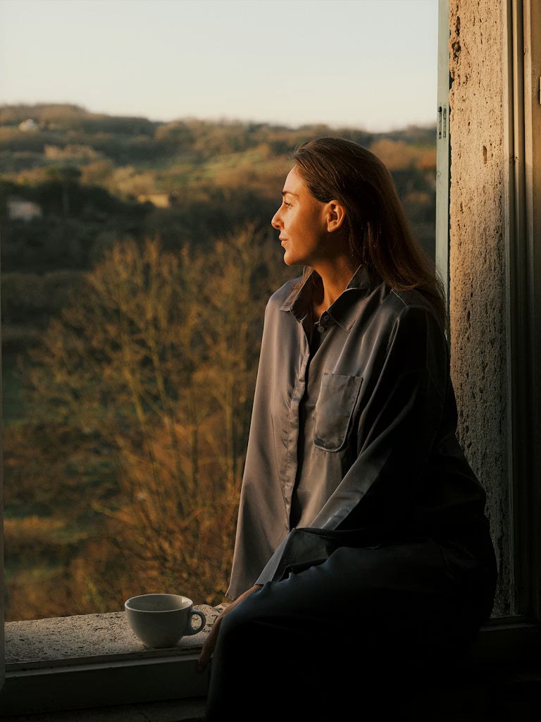 Woman seated beside a tall window looking toward a green landscape