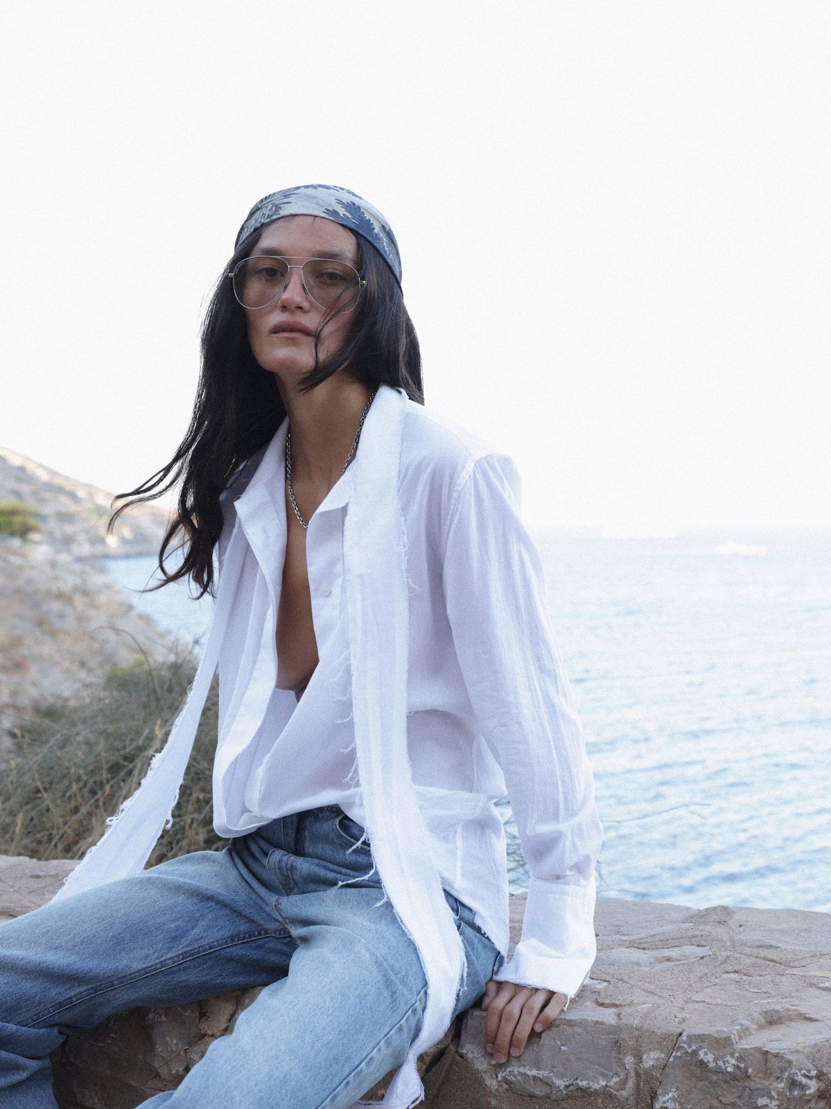 Model in a white shirt and faded jeans seated on a rocky shoreline