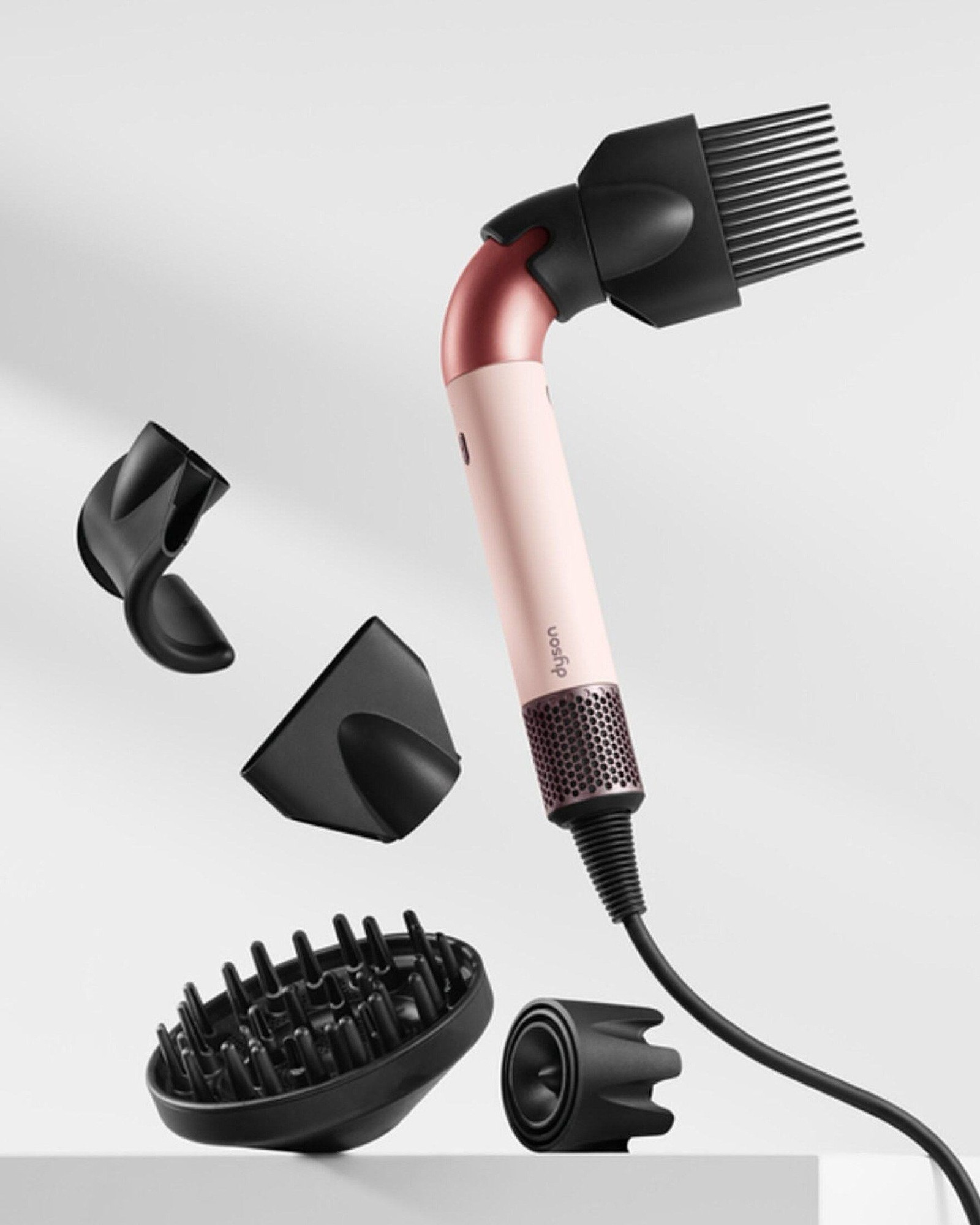 Pale pink and rose gold Dyson hair dryer with a curved neck and wide-tooth comb attachment, floating beside four black attachments on a white background