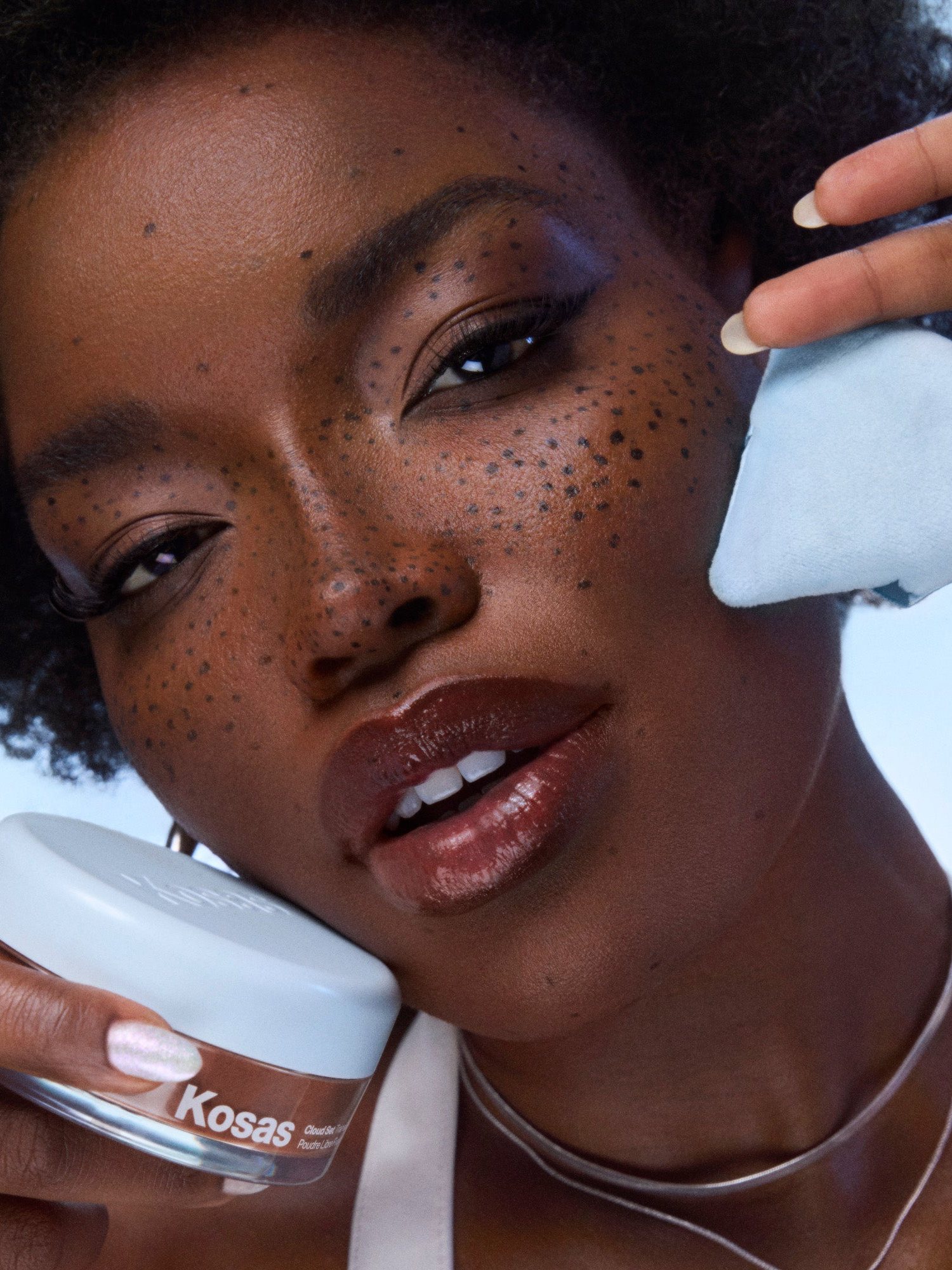 Model with freckles holding a Kosas Cloud Set powder jar and a blue puff against blue sky
