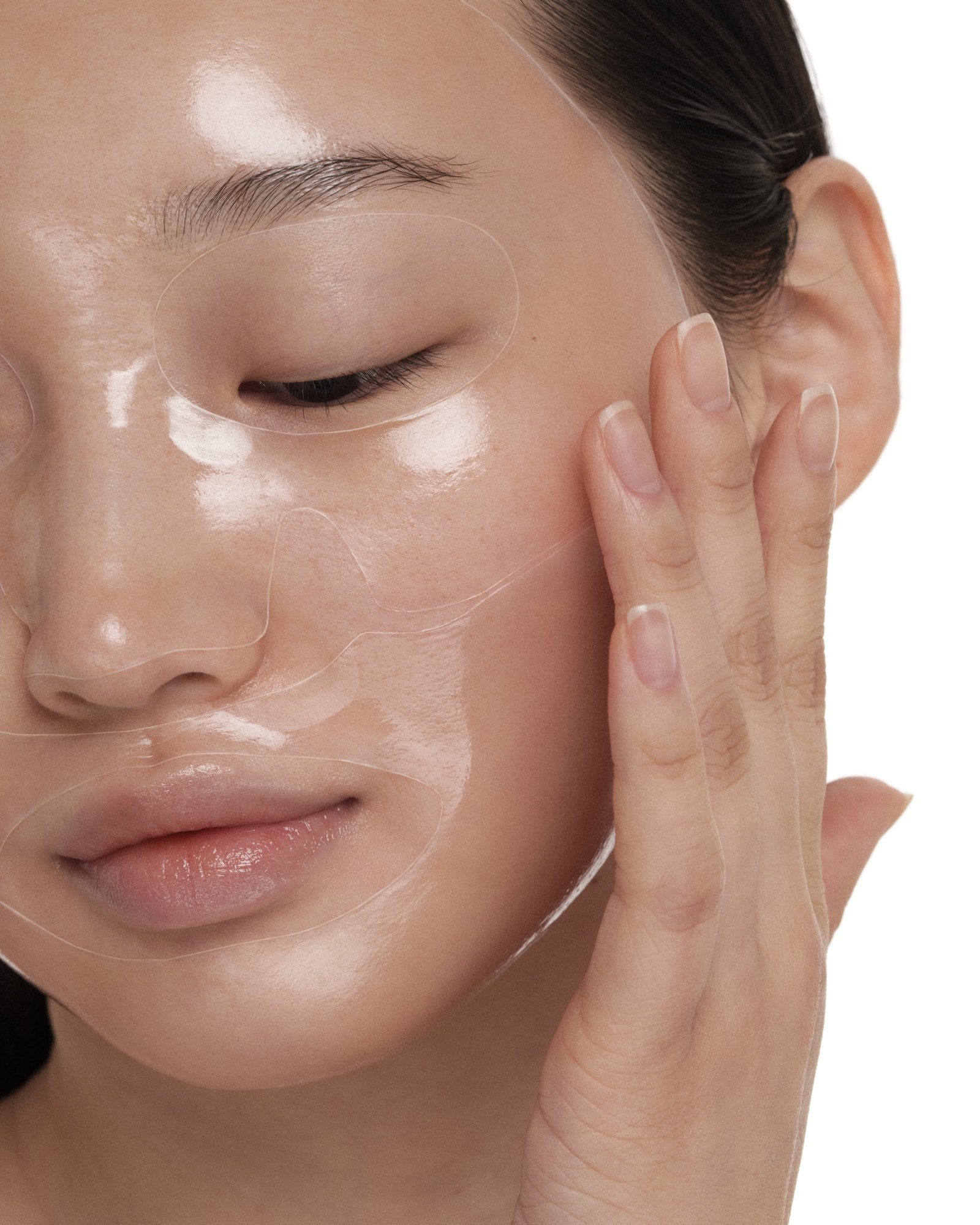 Close-up of a woman's glossy face wearing a clear hydrogel mask with eye and mouth cut-outs while she presses her cheek with her fingertips