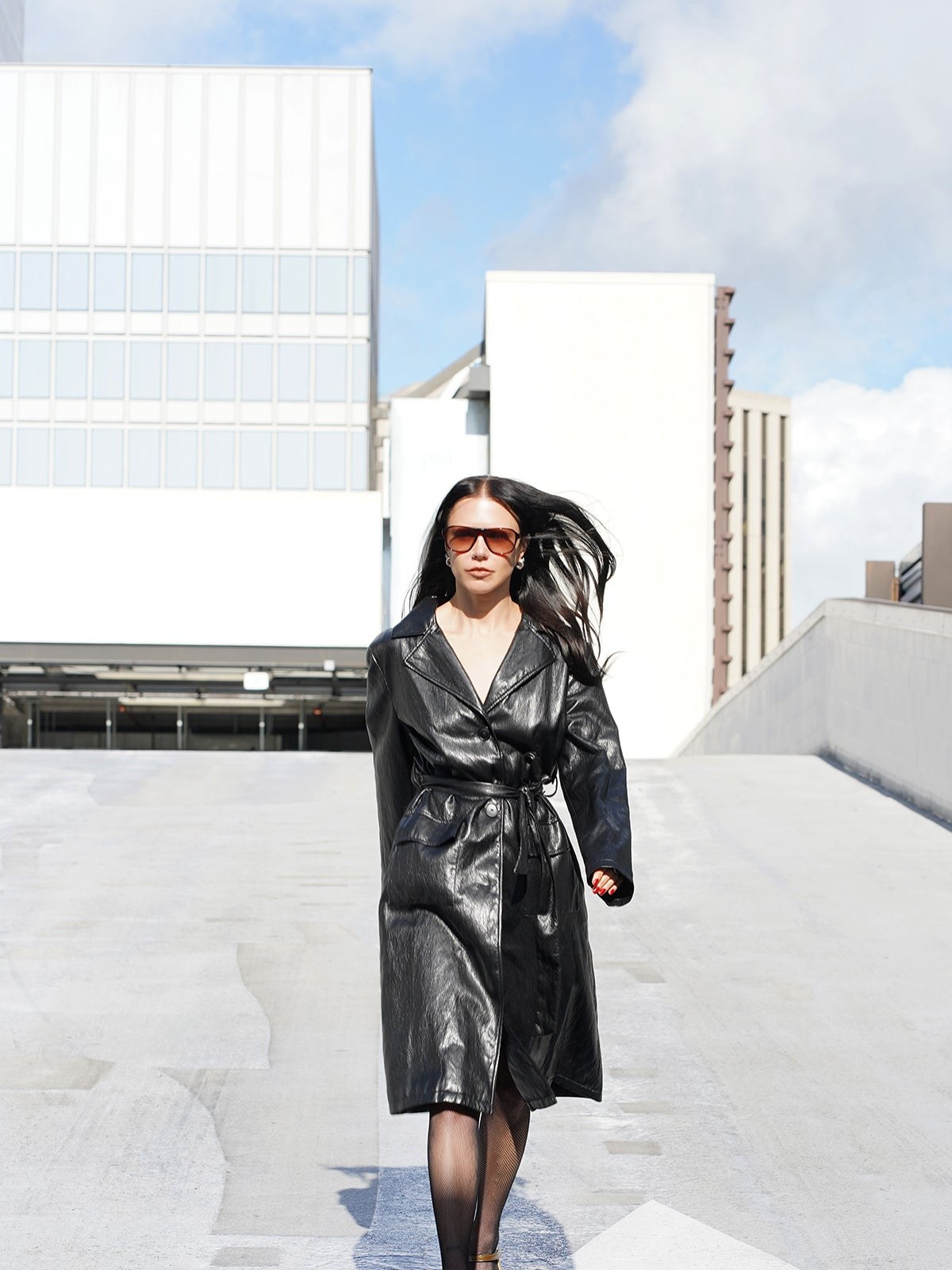 Model in a black leather-look coat walking across a bright city rooftop