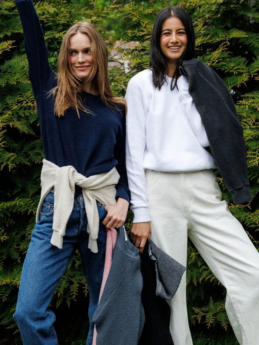 Two women in cozy tops and relaxed pants standing against leafy greenery