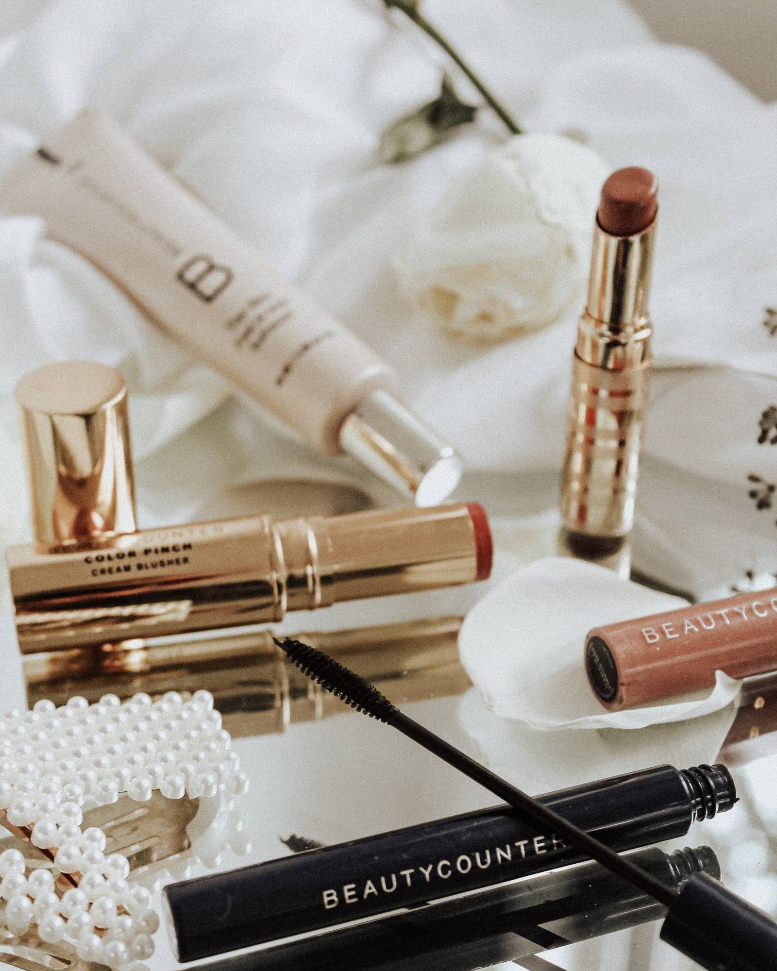 Gold cream blush, lipstick, a brow gel wand and a dark mascara tube on a mirrored tray beside white roses and a pearl hair clip