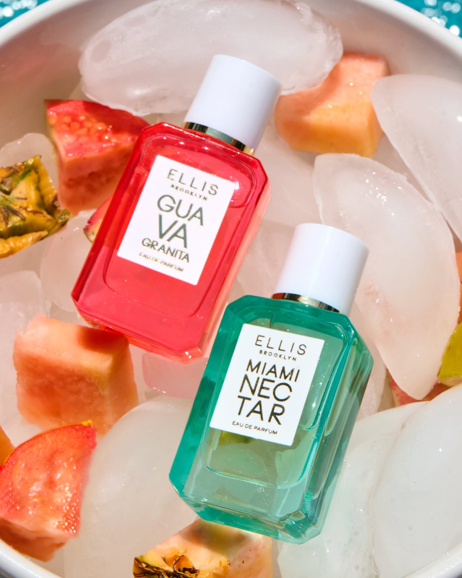 Two Ellis Brooklyn eau de parfum bottles, one pink and one aqua, resting on ice with cut guava and pineapple
