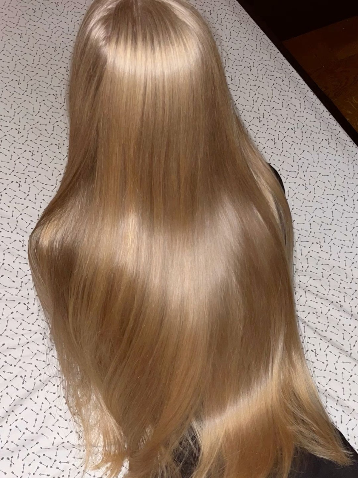 Back view of very long, smooth, glossy honey-blonde hair lying flat on a white patterned bedsheet