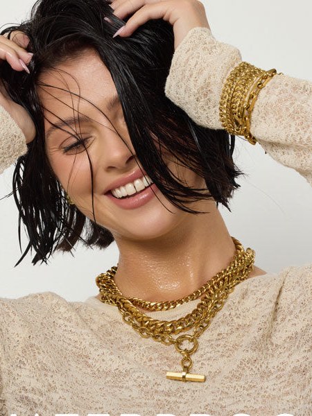 Smiling woman in a textured cream top wearing layered gold necklaces in a studio portrait