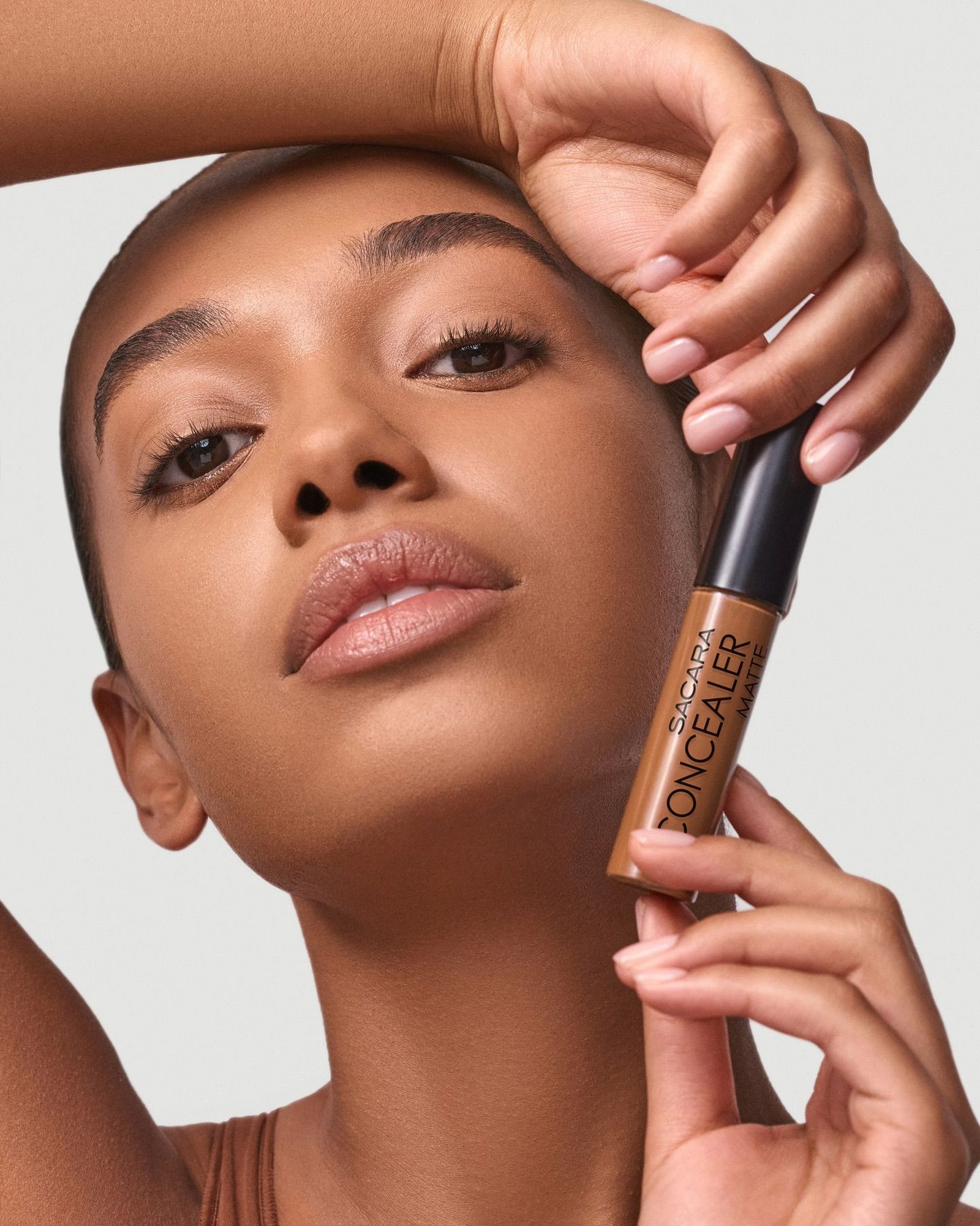 Model with a shaved head and brown eyes resting her arm on her head and holding a tube of matte concealer beside her face