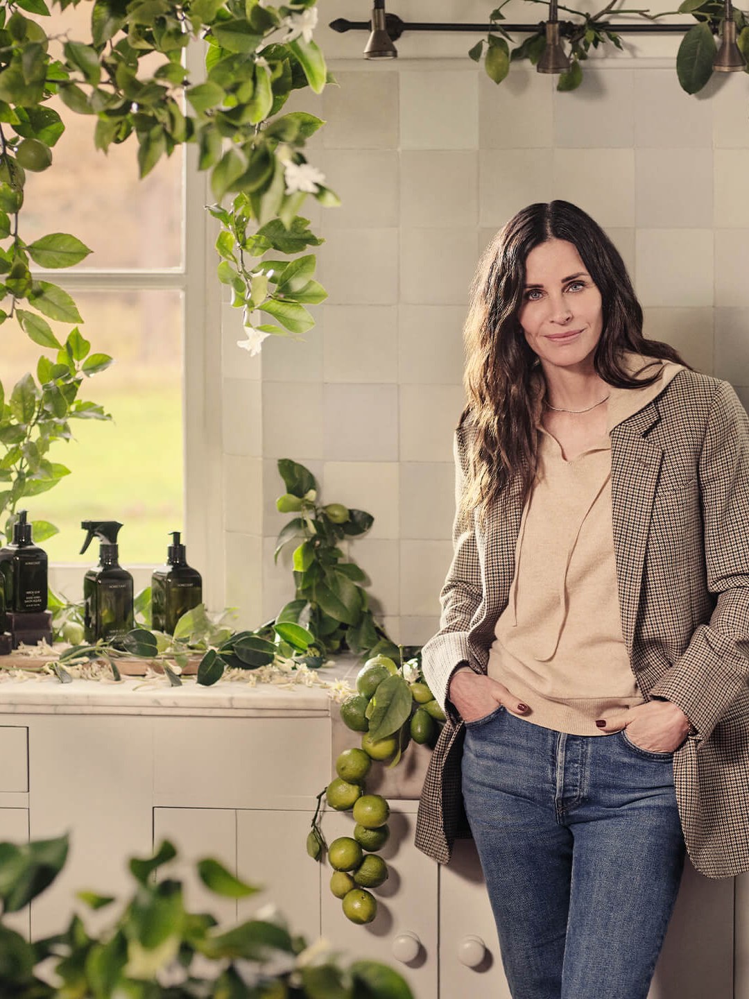 Courteney Cox beside Homecourt bottles on a green, plant-filled kitchen counter