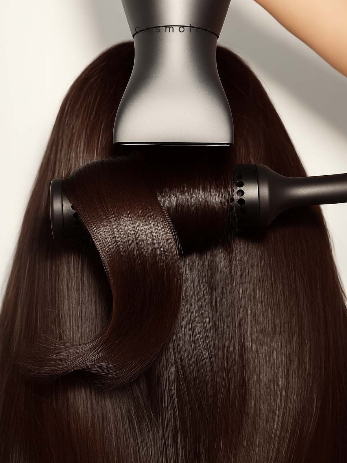 A silver hair dryer nozzle drying long dark brown straight hair wrapped around a black round brush