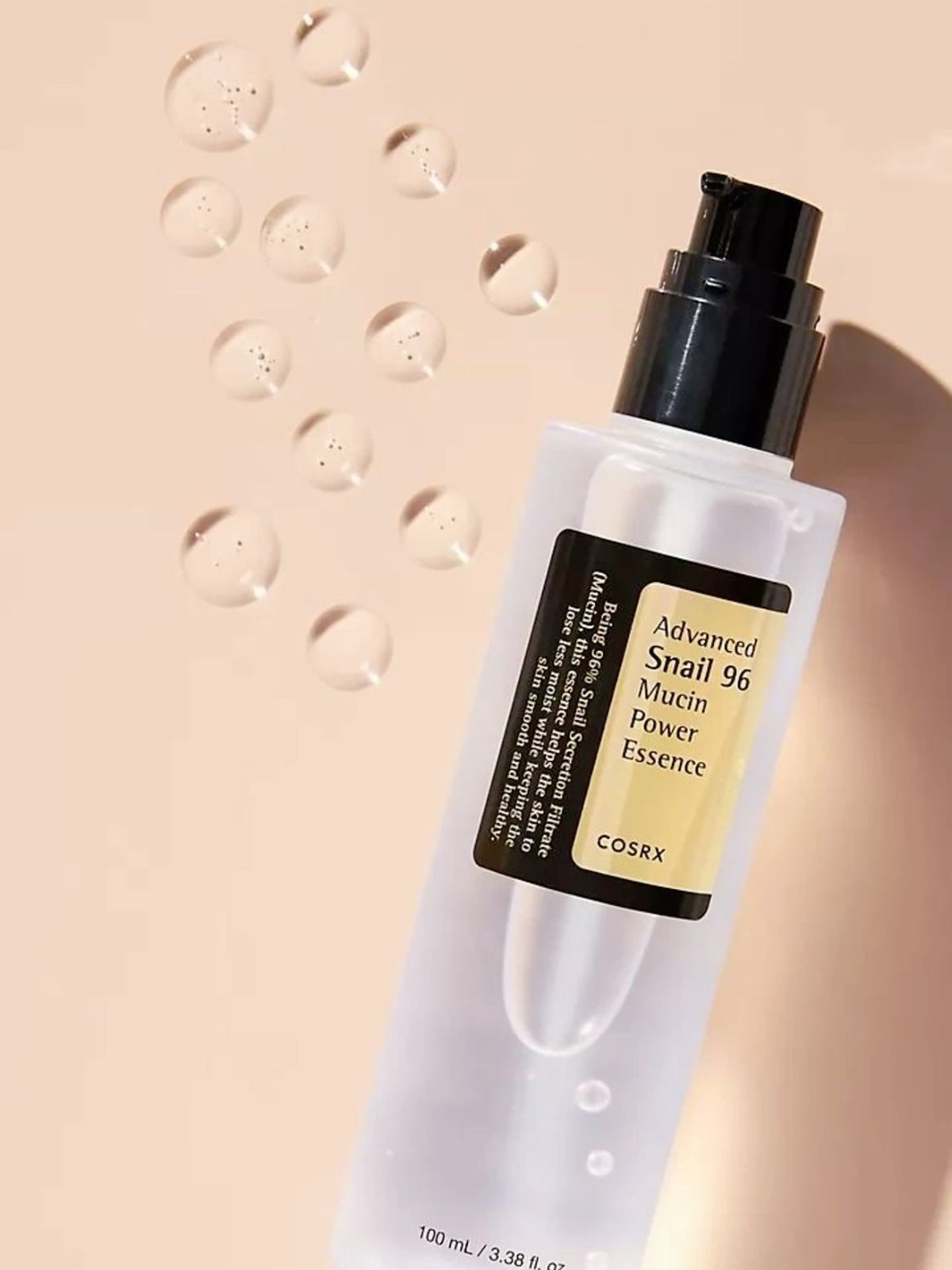 COSRX Advanced Snail 96 Mucin Power Essence bottle lying on a peach background beside rows of clear droplets