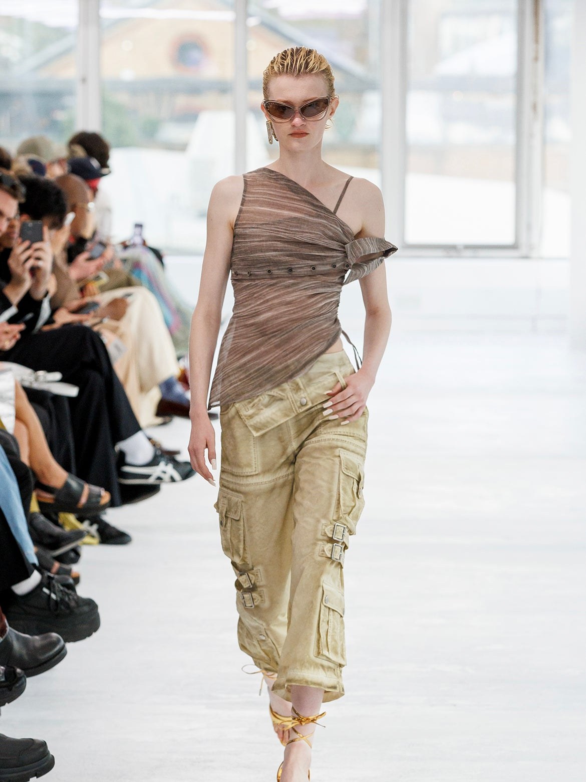 Model in a draped taupe top and wide trousers on the runway