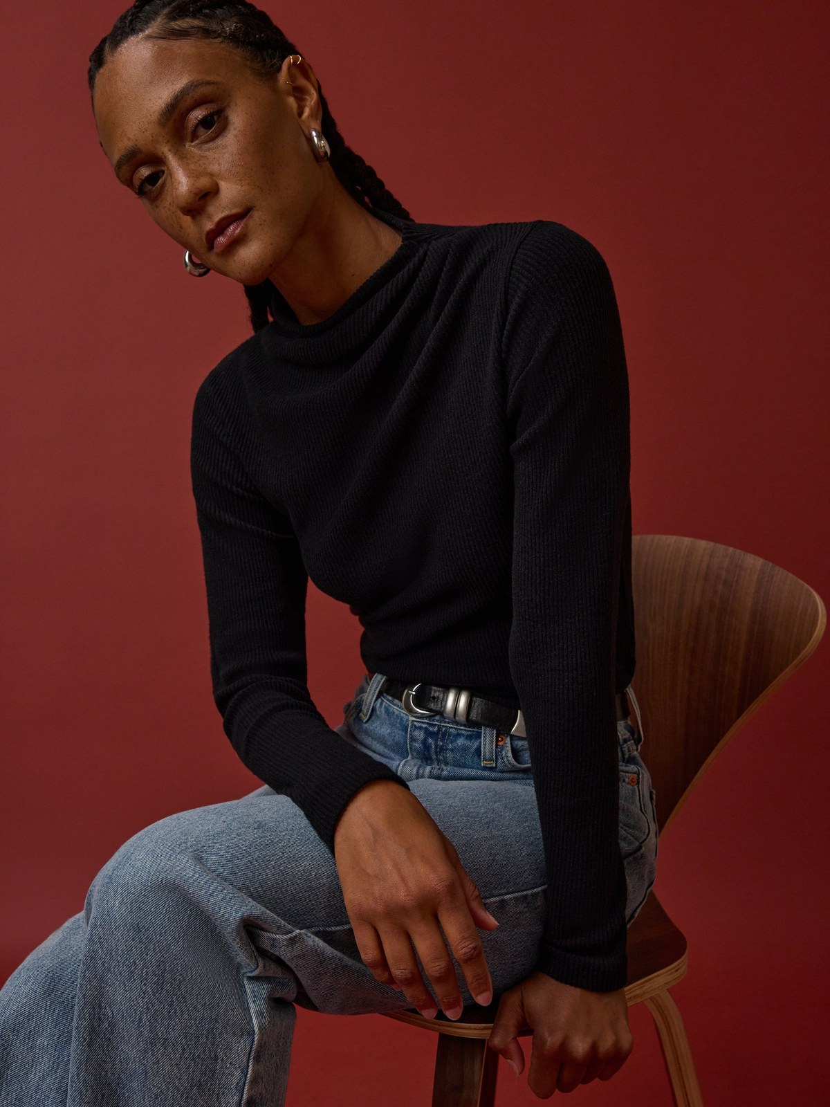 Model in a black top and jeans seated on a chair against a red backdrop