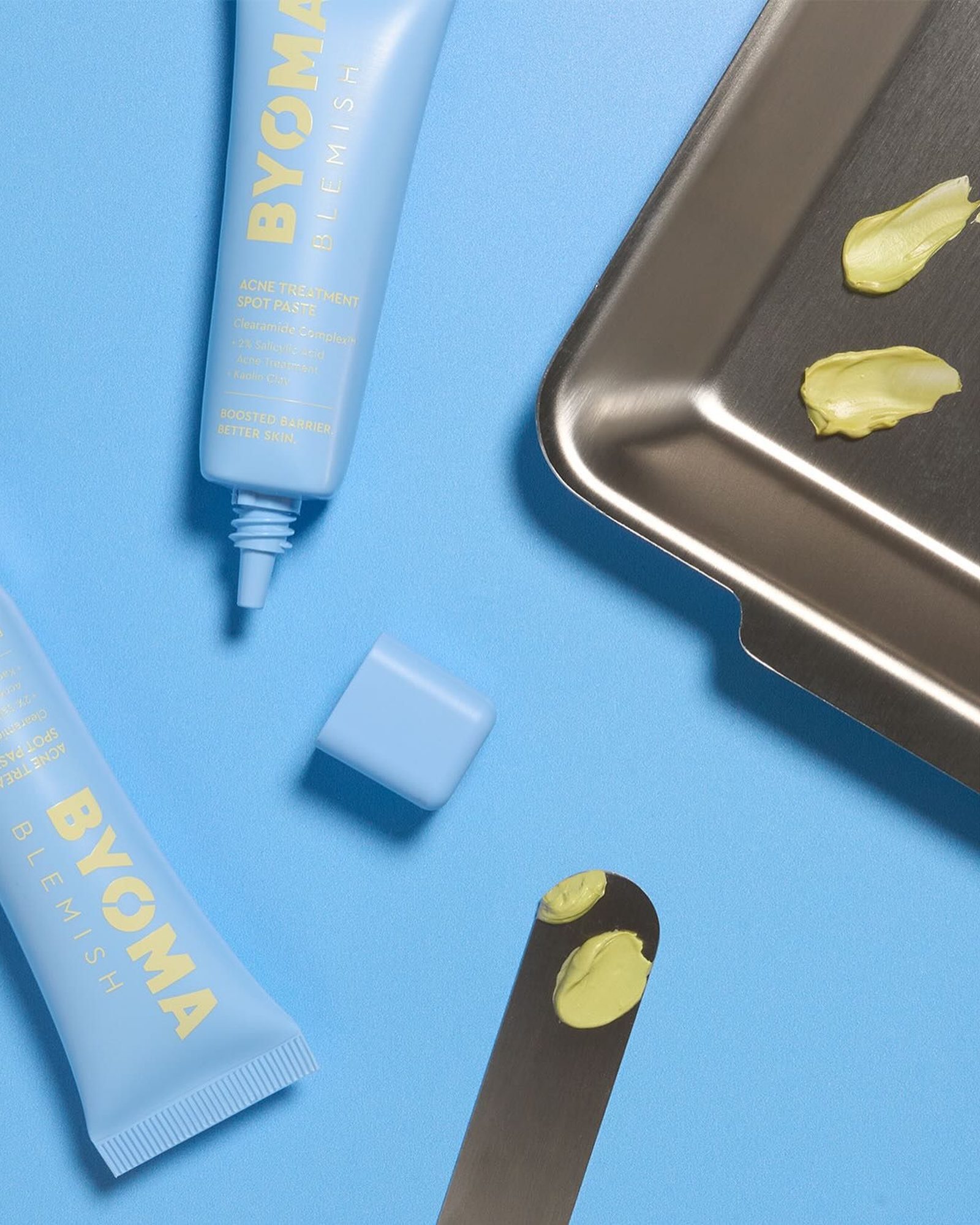 Light blue BYOMA Blemish Acne Treatment Spot Paste tubes on a blue background with swipes of pale green paste on a metal tray and spatula