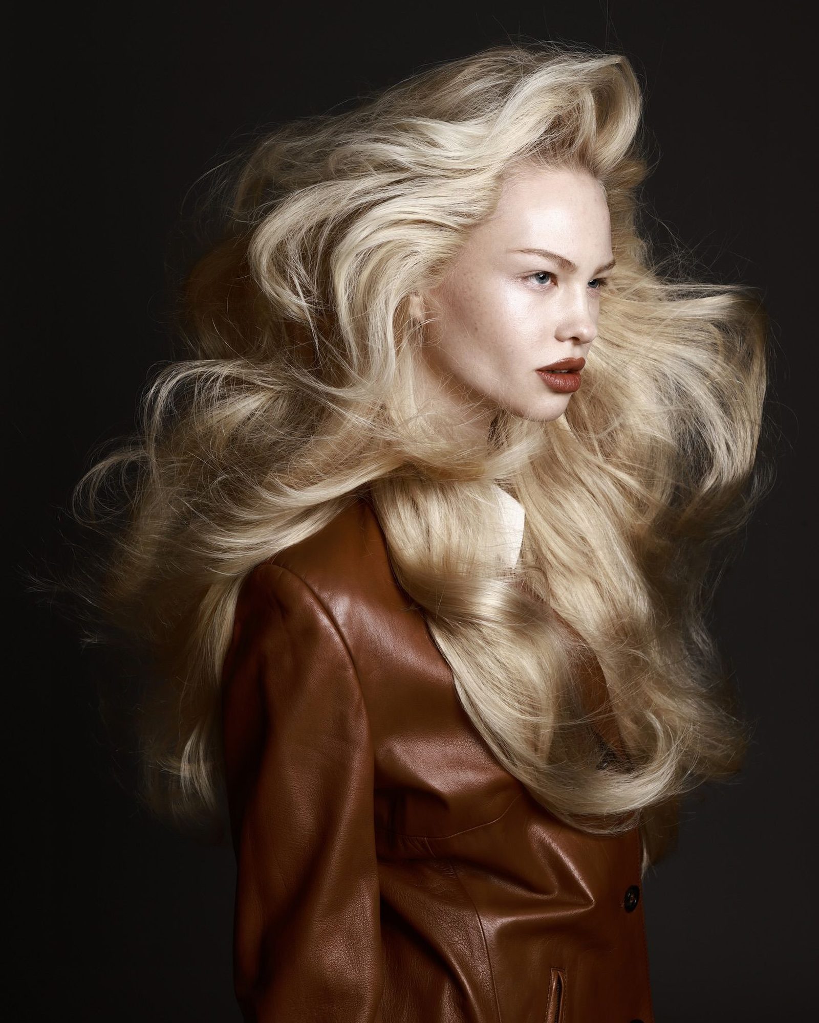 Woman with a voluminous platinum blonde blowout in a brown leather coat against a dark backdrop