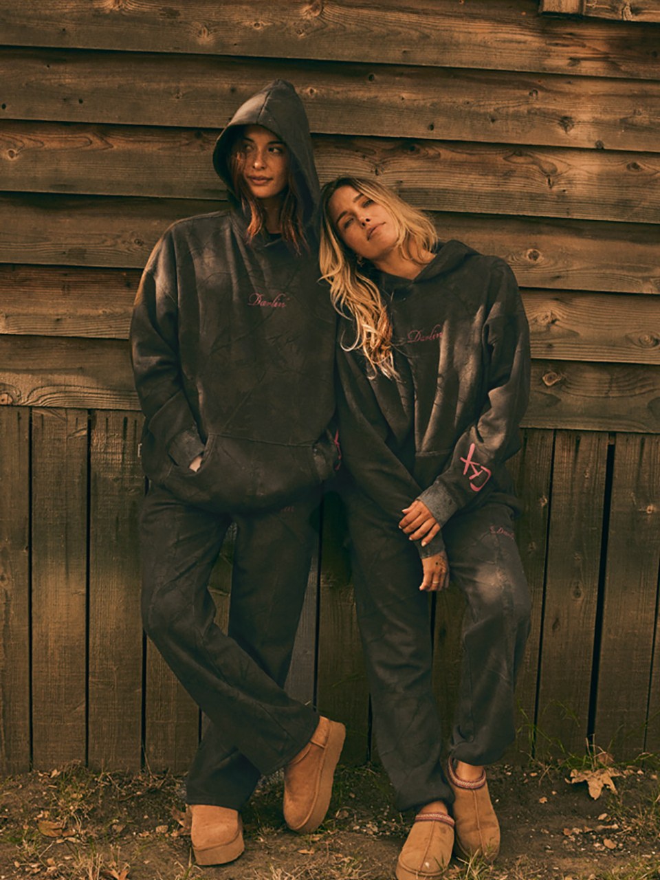Two women in oversized western sweatshirts standing by a wooden barn wall