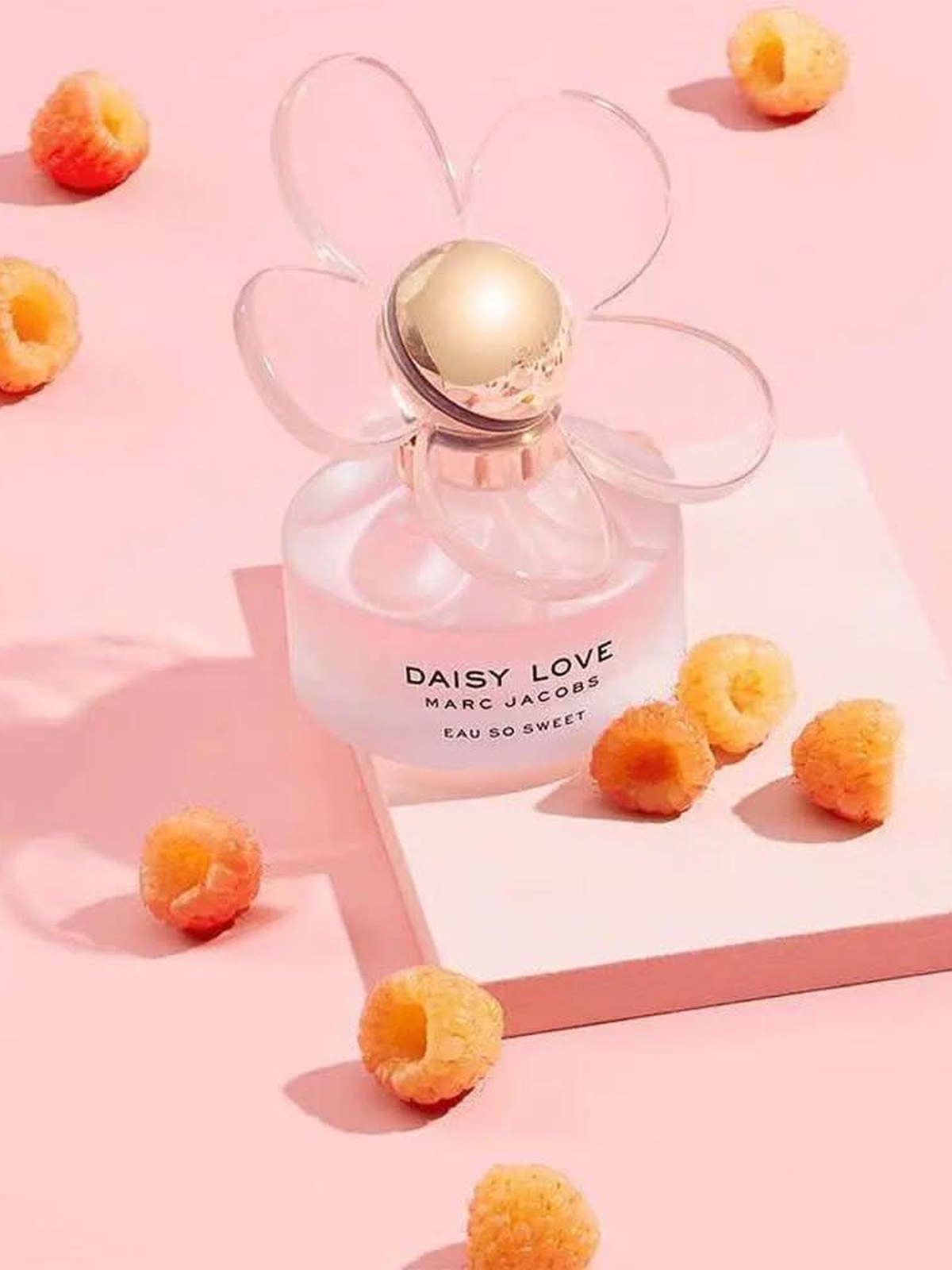 Daisy Love Eau So Sweet bottle with a clear flower cap on a pale pink block surrounded by golden raspberries