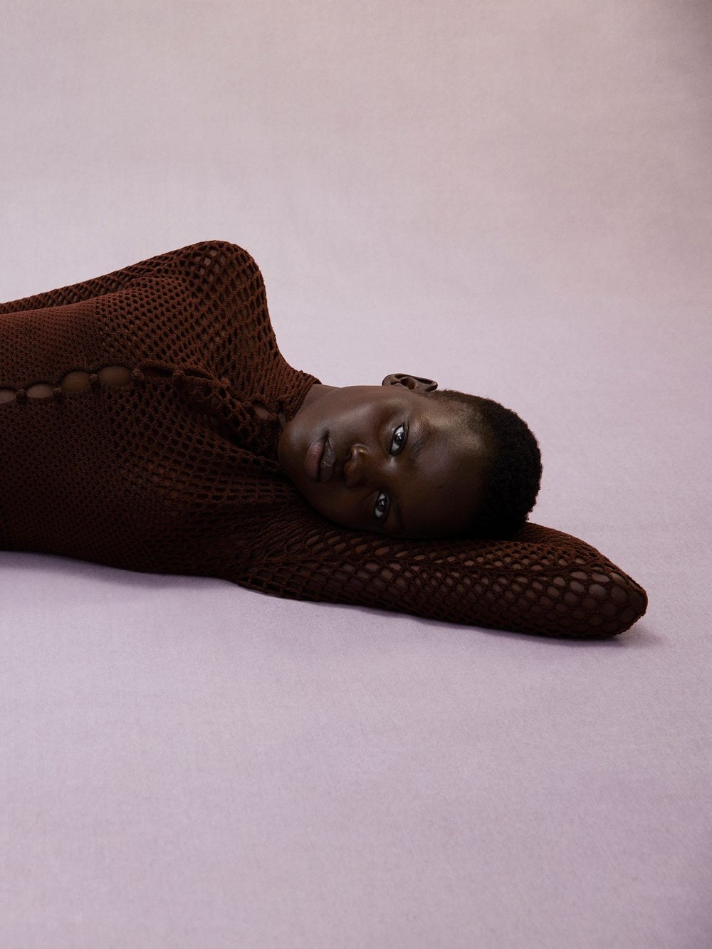 Model in a dark open-knit set reclining against a lavender studio backdrop
