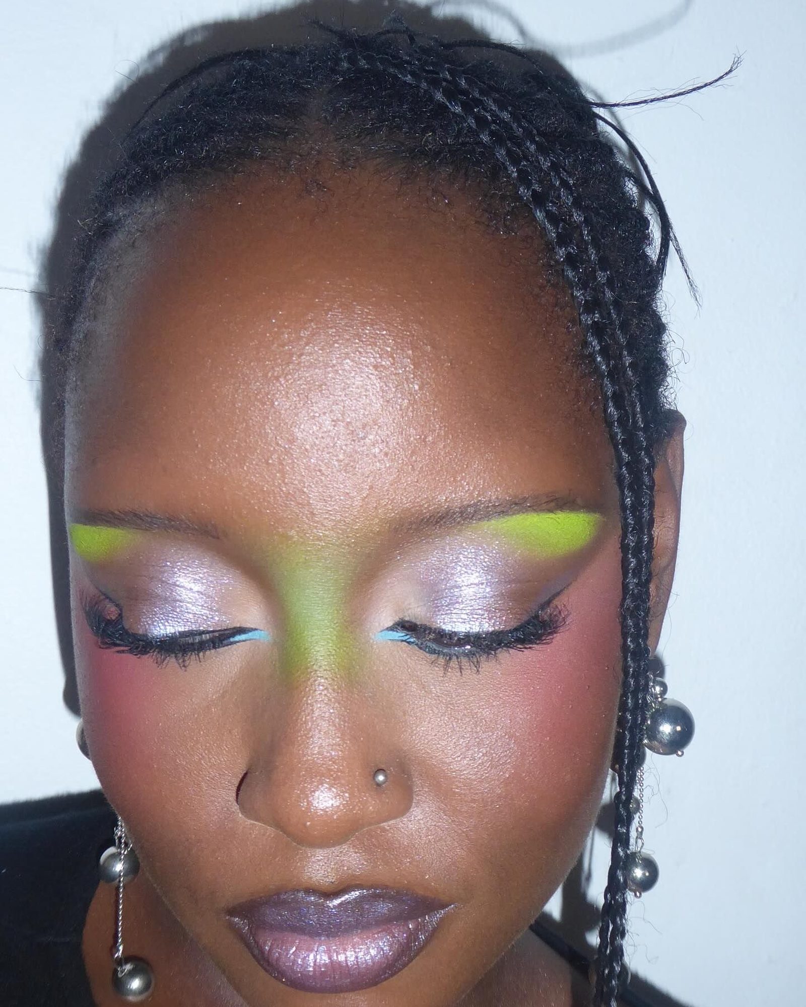 Close-up of a woman with braids looking down, wearing lime green and lilac shimmer eyeshadow with blue liner, pink blush and a mauve lip, silver ball earrings