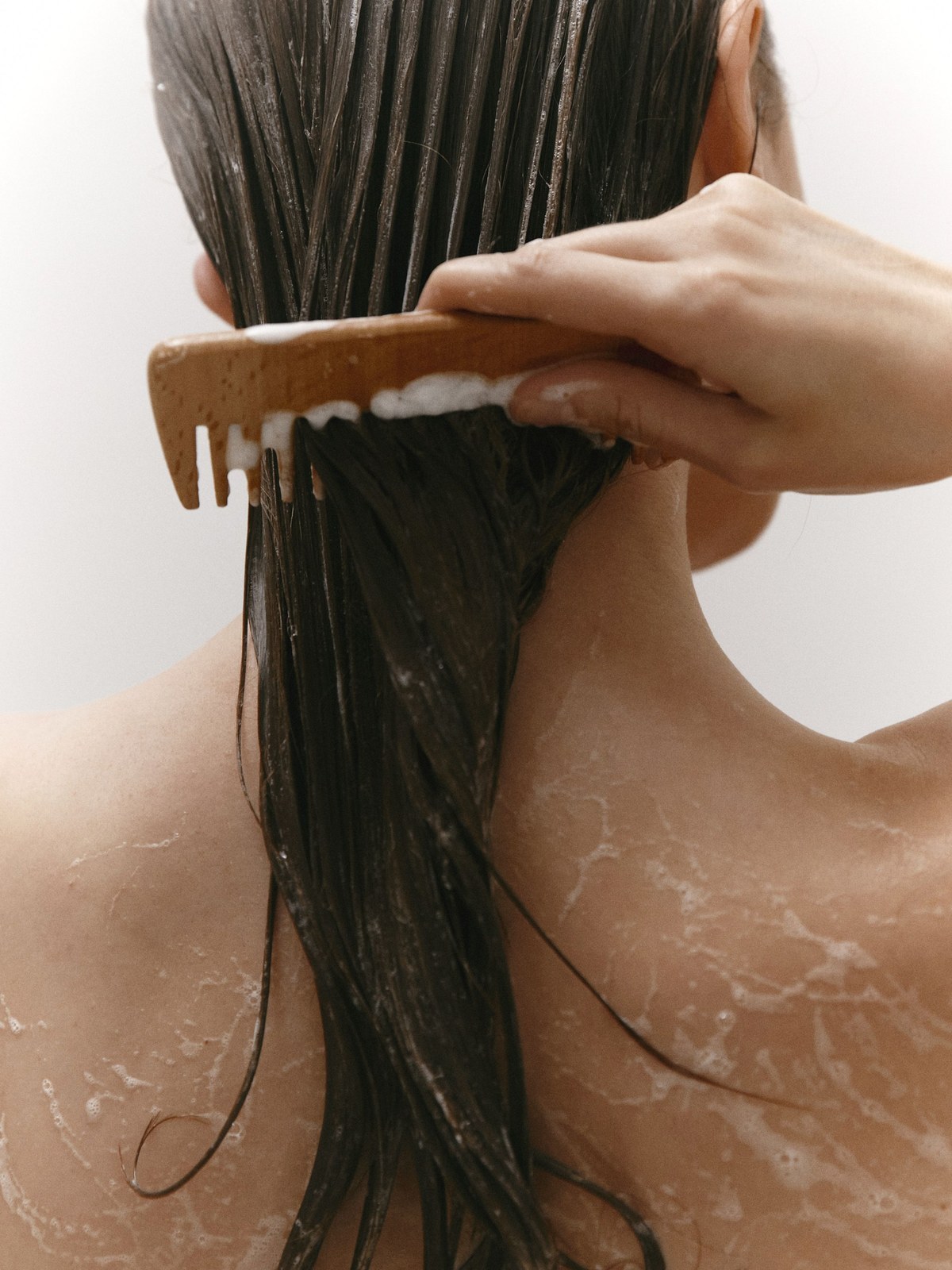 Back view of a woman in the shower running a wooden wide-tooth comb through long wet brown hair, with suds on her shoulders