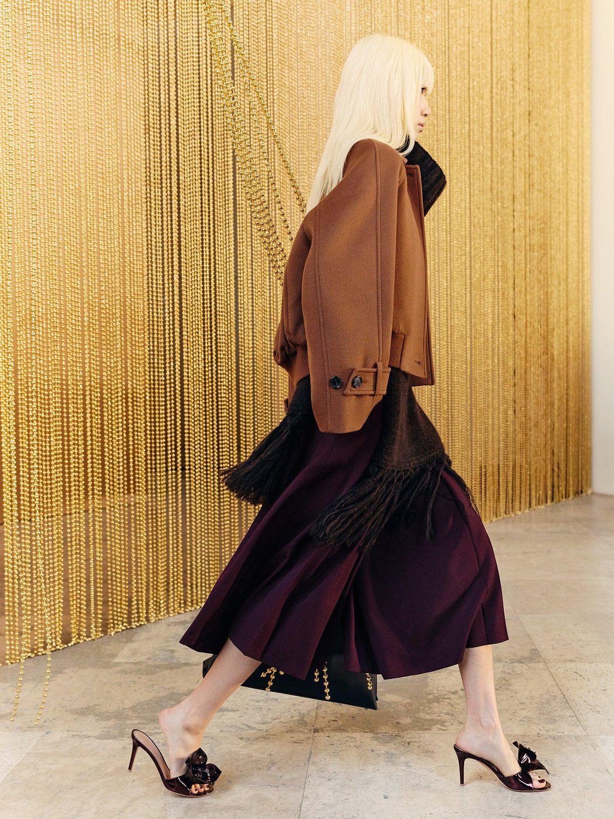 Model in profile in a brown jacket and plum skirt walking past a gold chain curtain