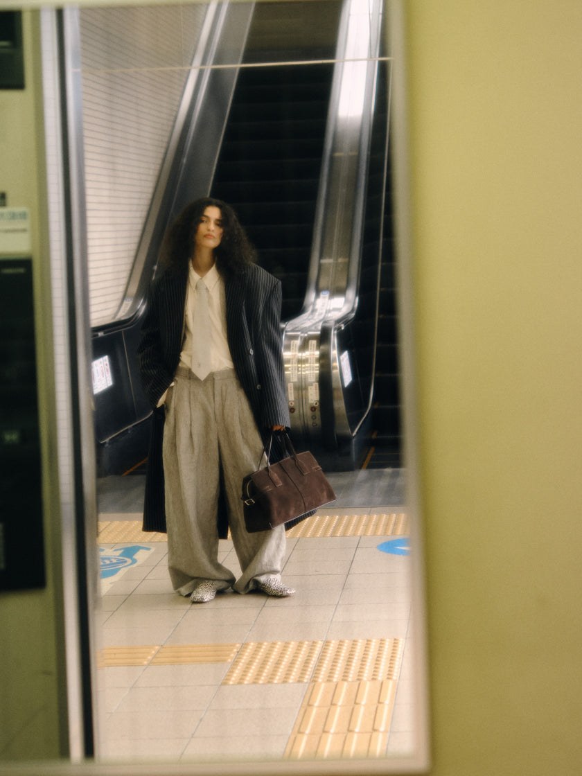 Model carrying a brown bag on a station platform beside a train