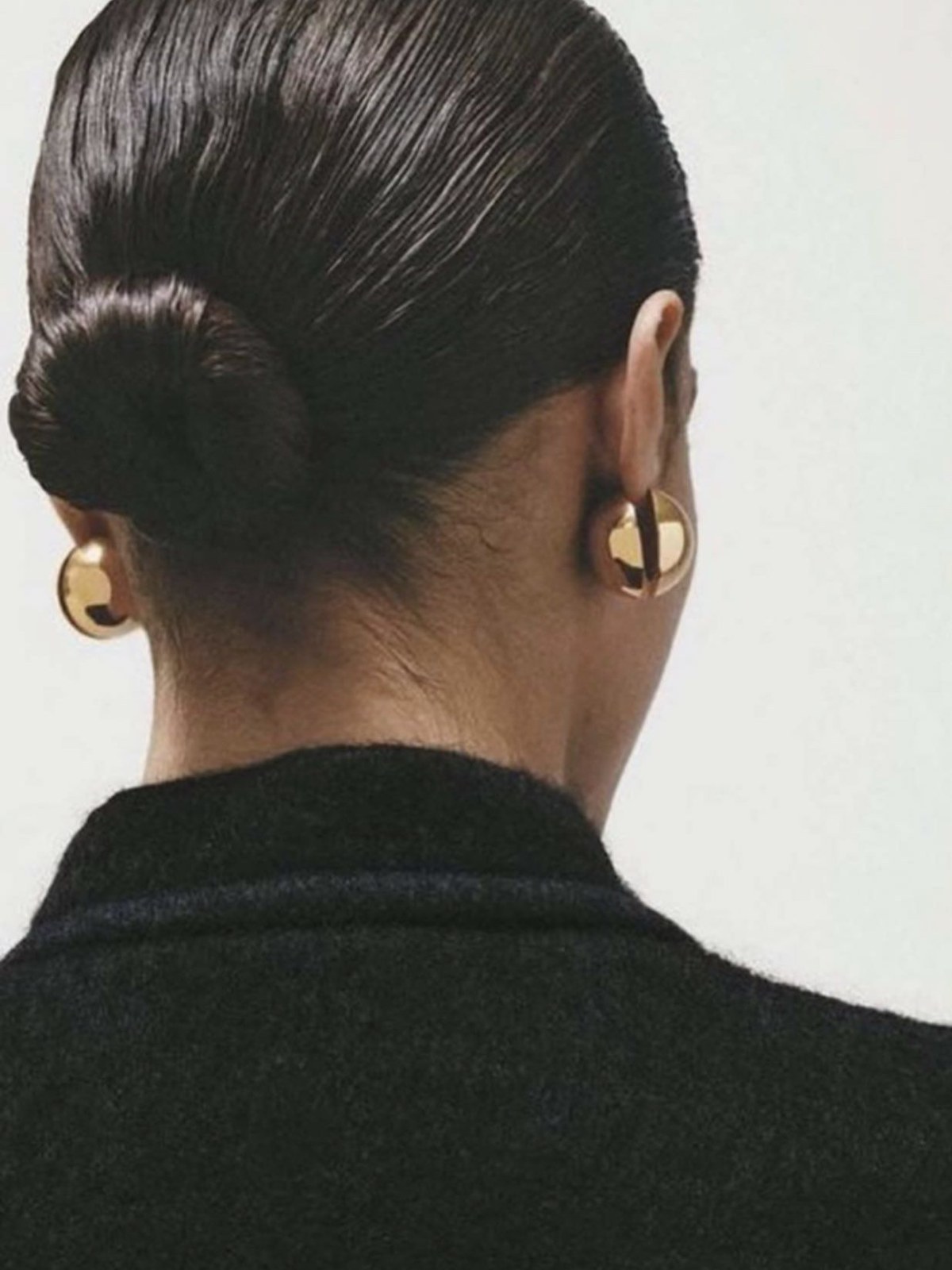 Back view of a woman with wet-look slicked-back dark hair in a low bun, wearing large gold dome earrings and a black knit collar