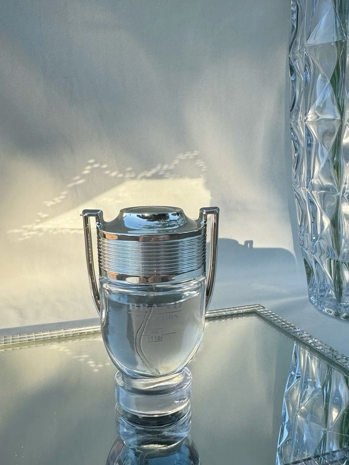 Silver trophy-shaped perfume bottle on a mirrored tray beside a faceted crystal vase