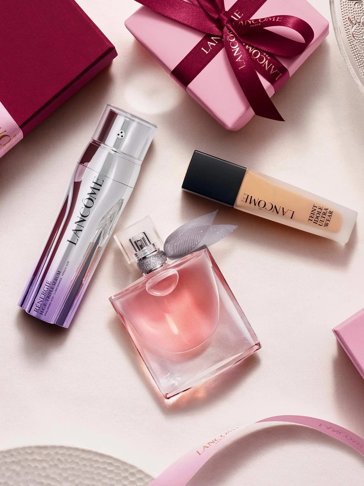 Flat lay of a serum, foundation and La Vie Est Belle perfume on cream linen with pink gift boxes
