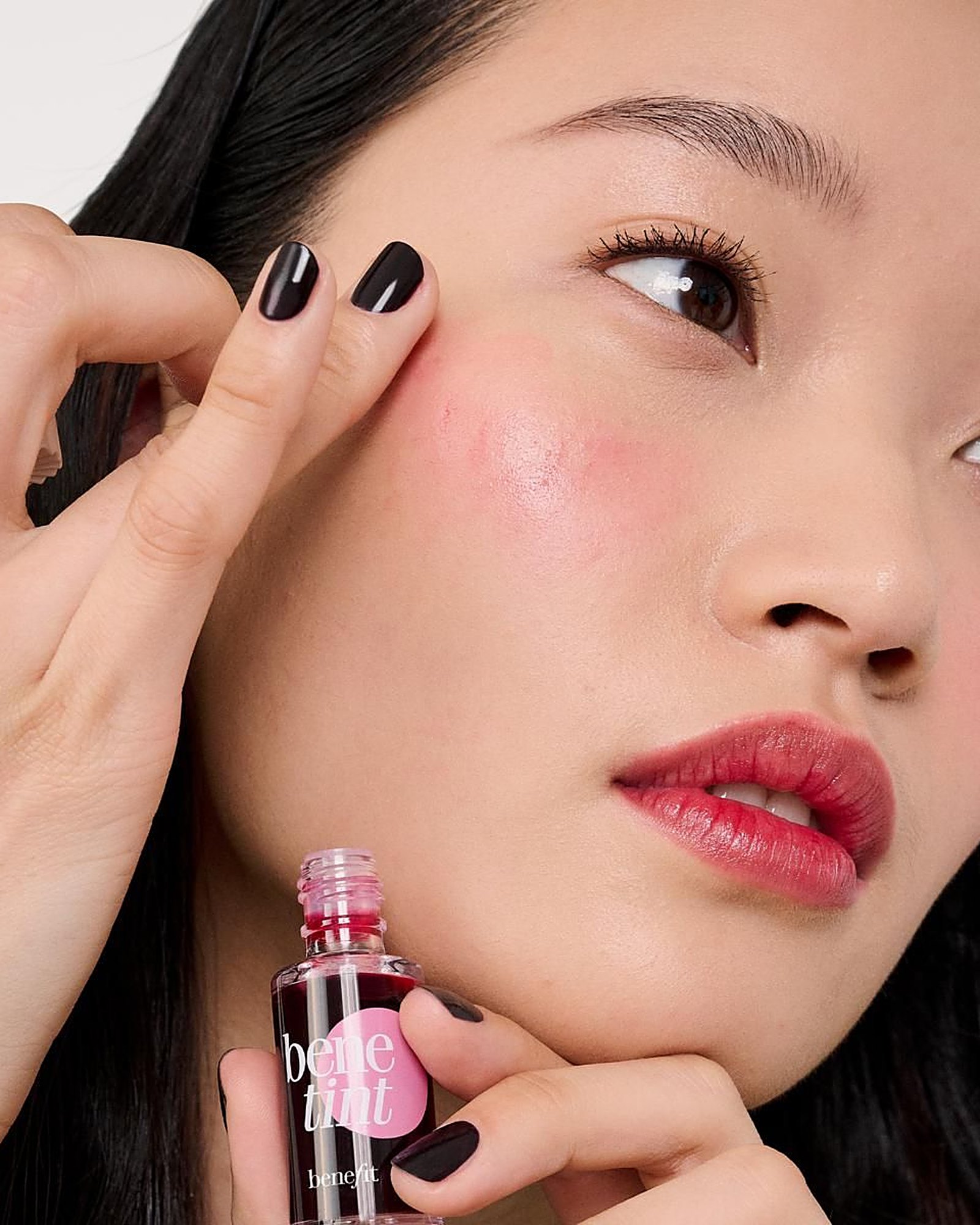 Close-up of a woman with dark nails tapping a pink Benetint flush onto her cheekbone while holding the Benetint bottle