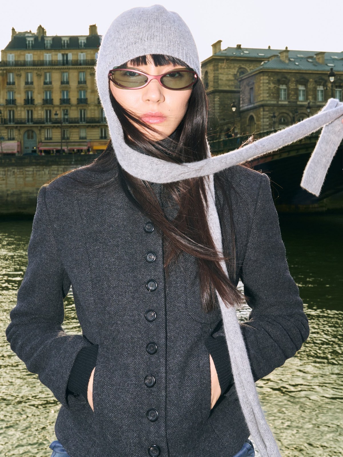 Woman in BRU sunglasses, a gray beanie and dark tailored jacket by the river