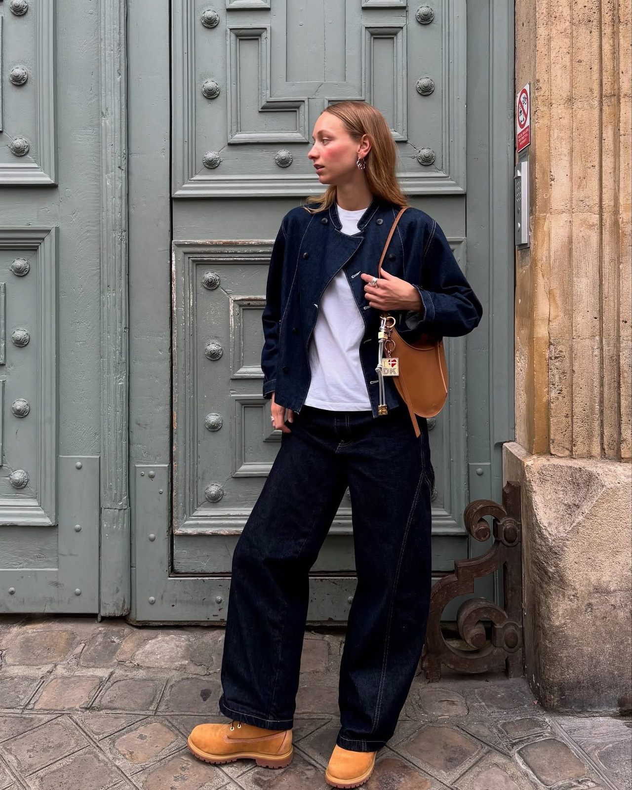 Woman by a grey door in a navy jacket and wide navy trousers with wheat Timberland boots and a tan bag