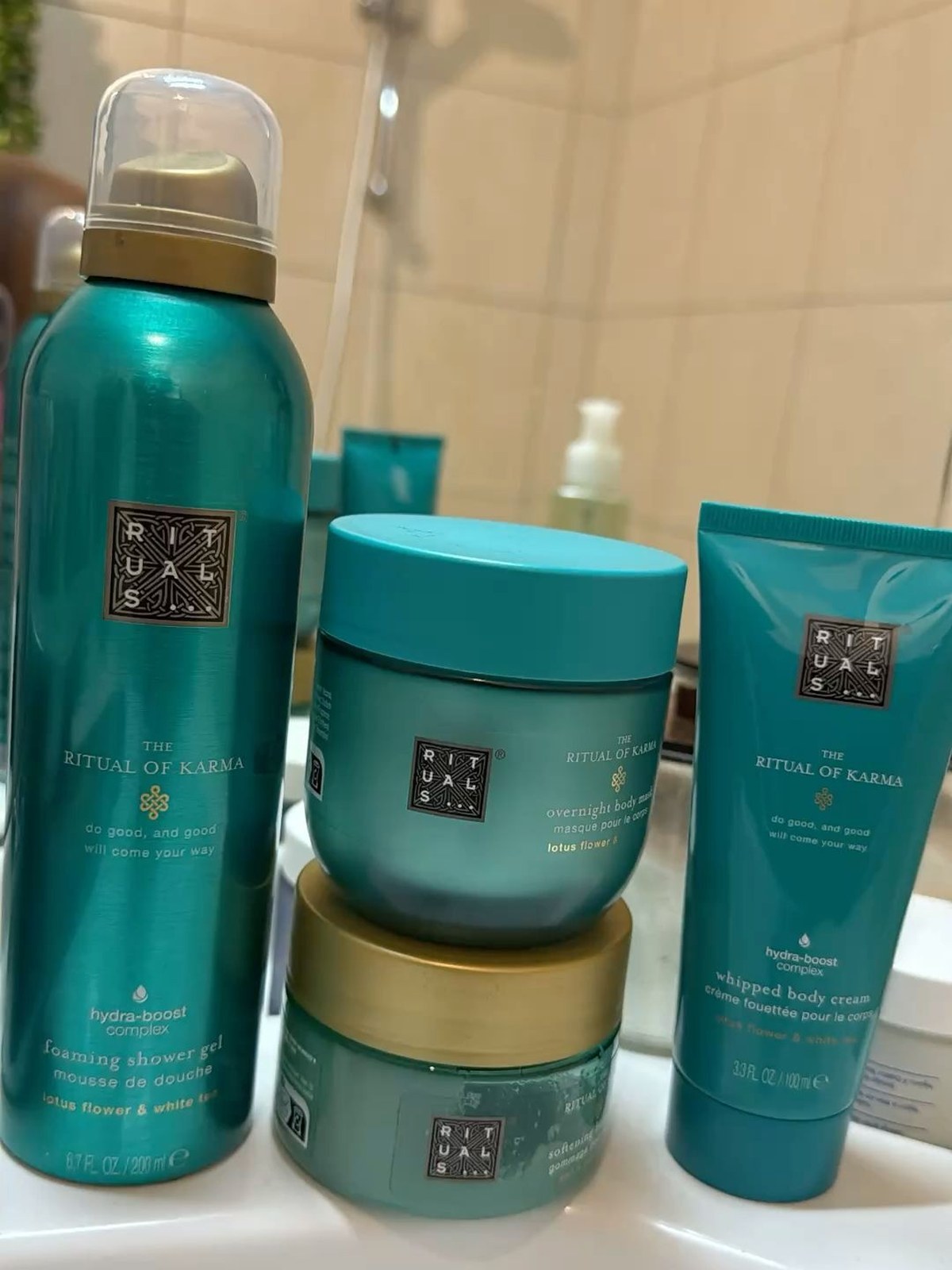 Bathroom shelf with teal The Ritual of Karma foaming shower gel, overnight body mask, softening scrub and whipped body cream beside a shower