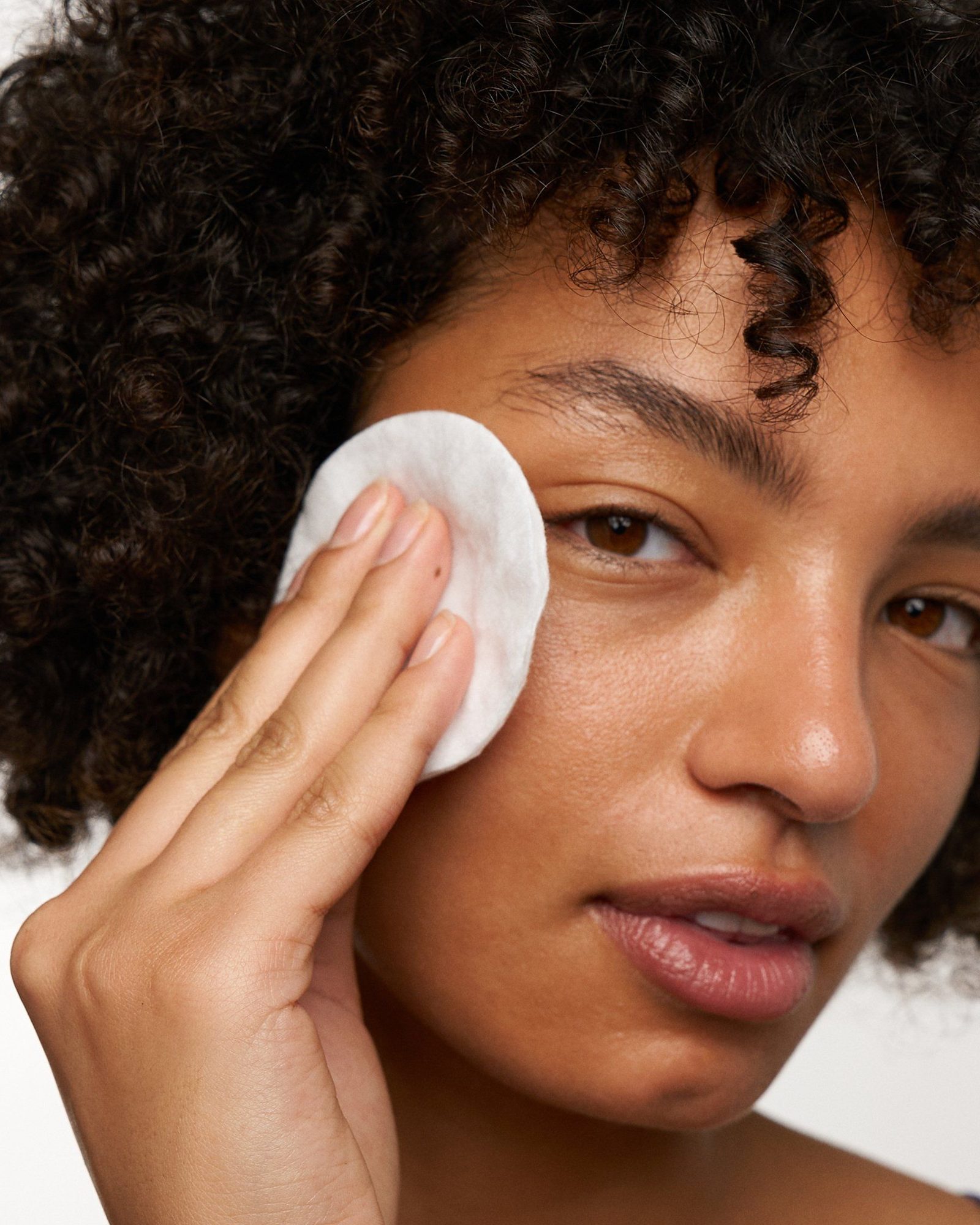 Close-up of a woman with dark curly hair and a glowing complexion pressing a round white cotton pad against her cheek