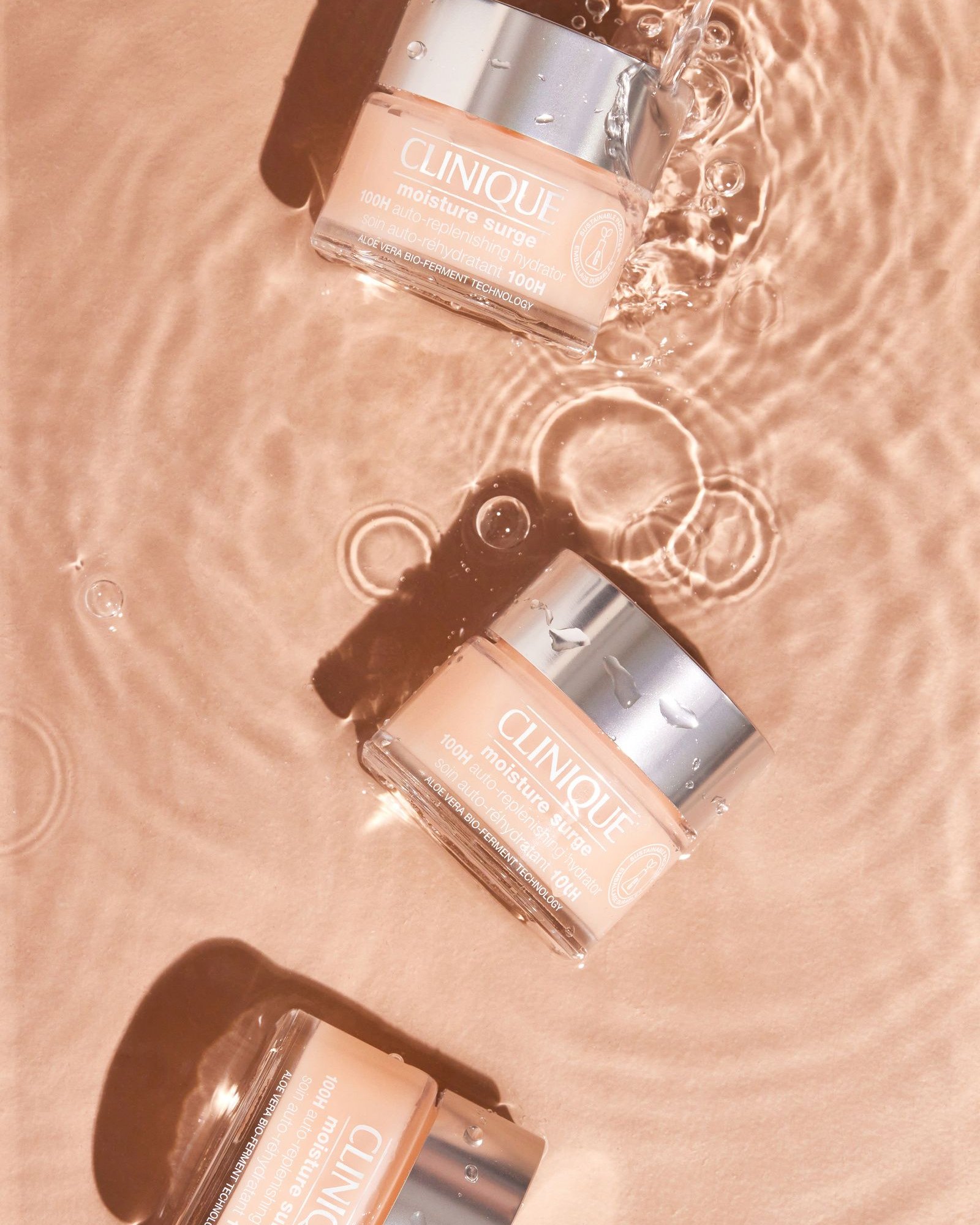 Overhead shot of three Clinique Moisture Surge 100H jars with silver lids floating in peach-tinted rippling water