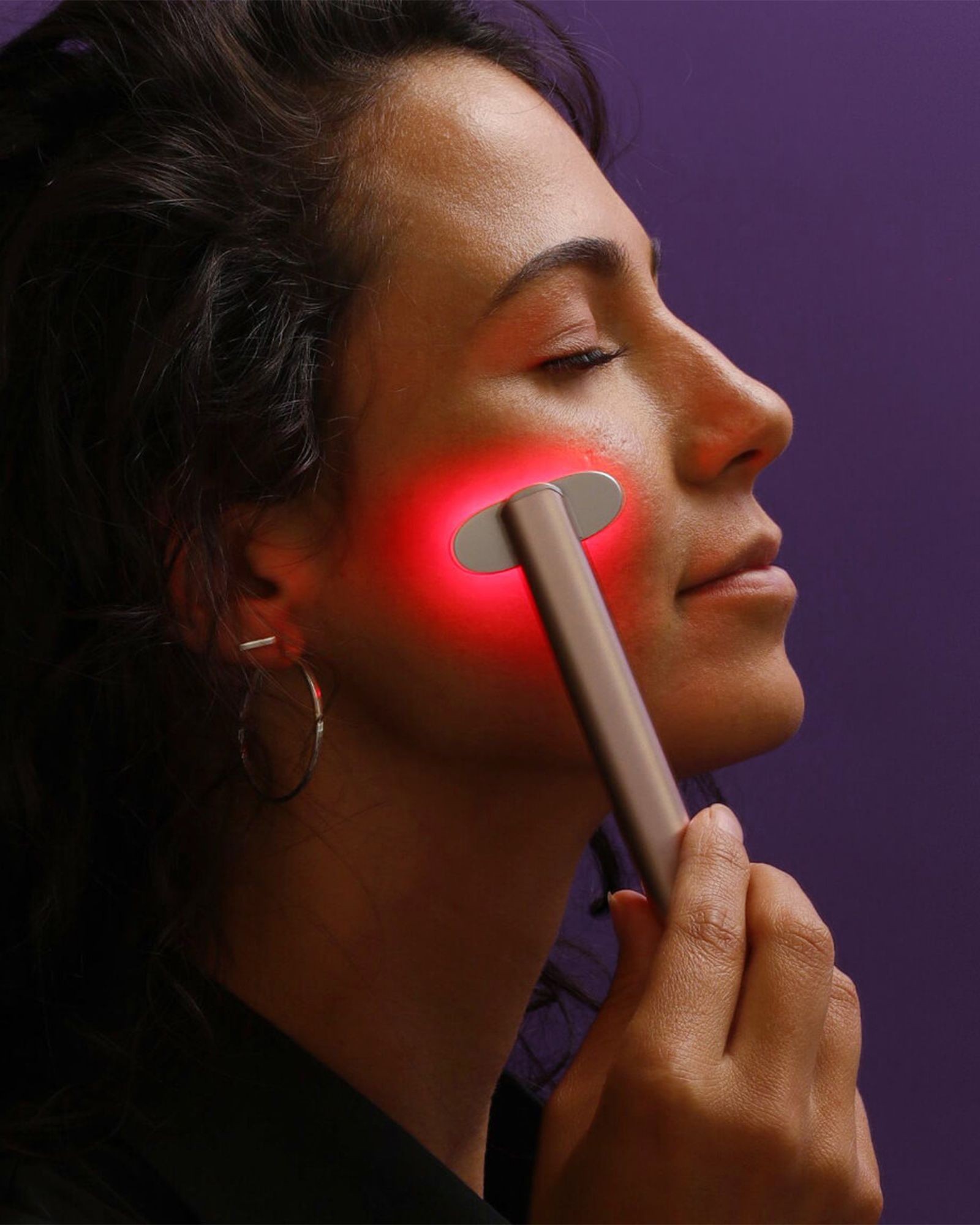 Close profile of a woman with dark hair holding a red light wand to her cheek against a purple wall