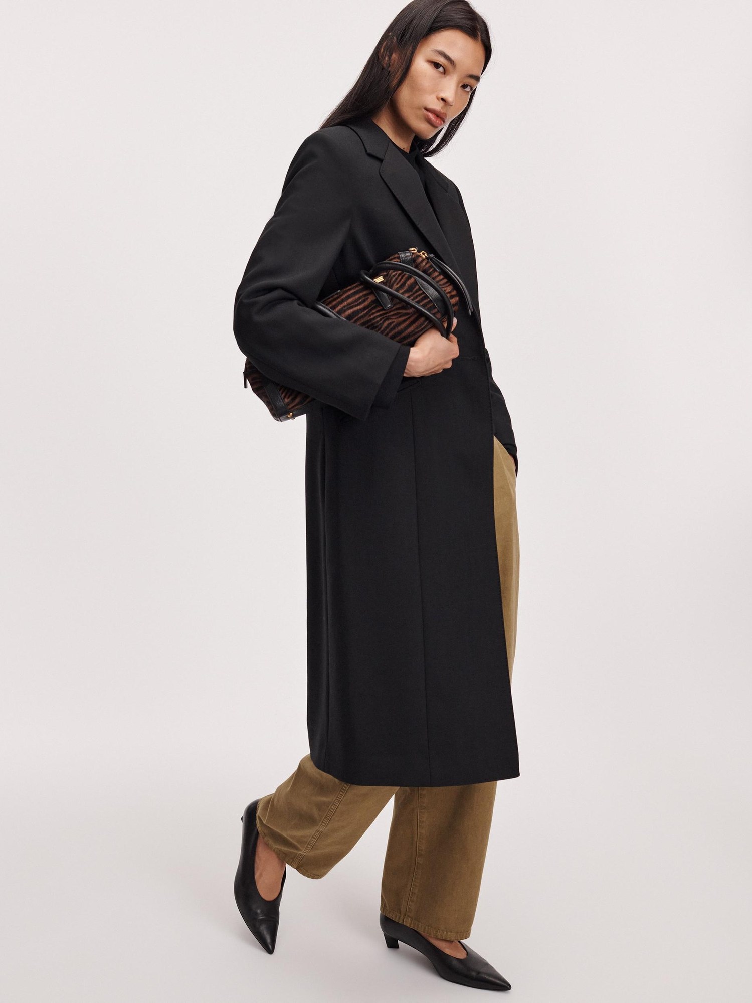 Rag & Bone model in a long black coat and khaki trousers carrying a brown bag