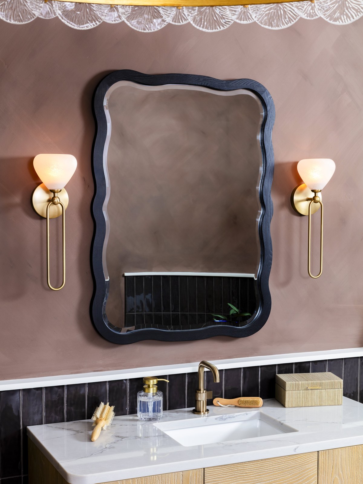 Wavy black framed mirror above a bathroom vanity between wall sconces