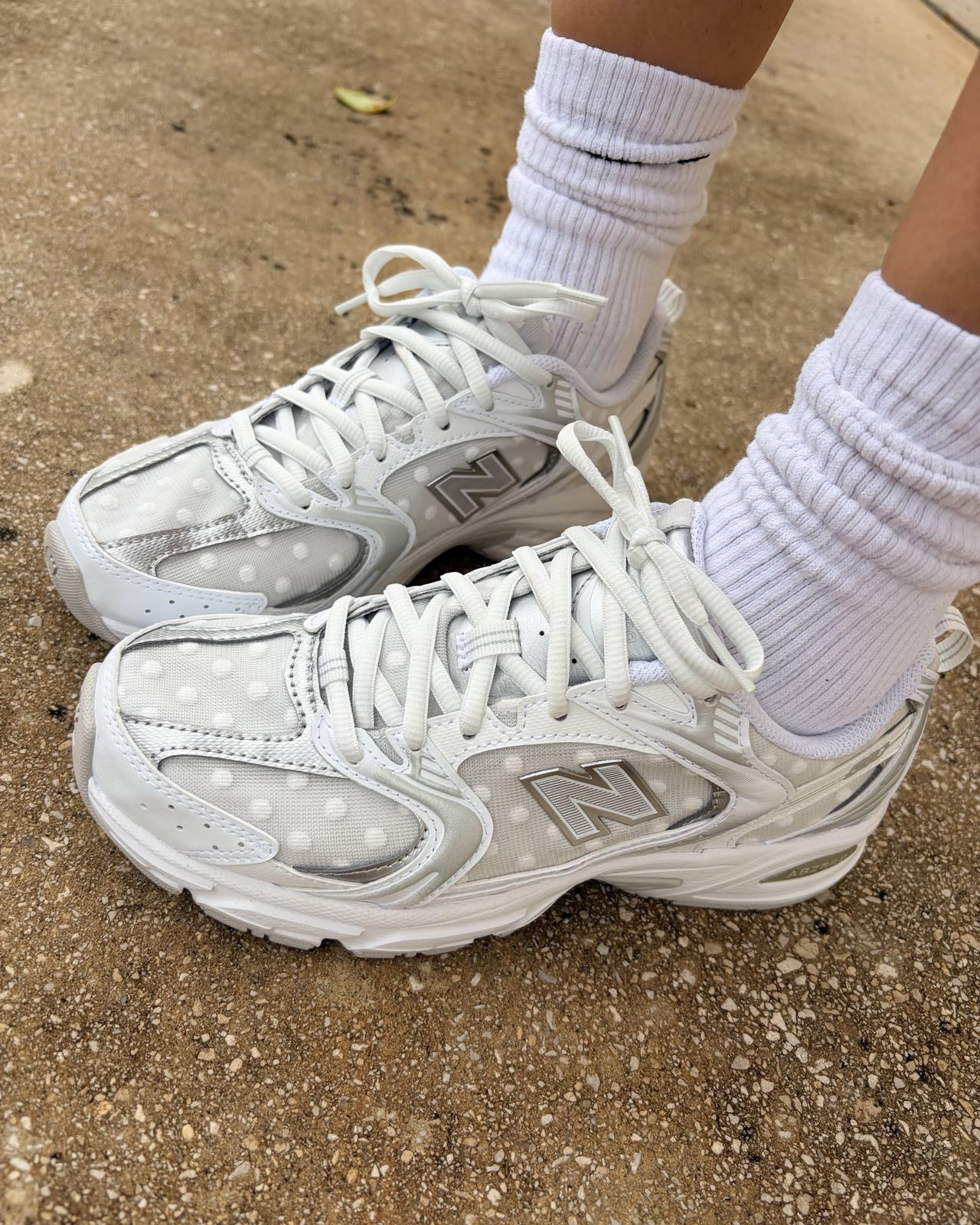 Close-up of feet in white socks and New Balance 530 sneakers with a silver polka dot mesh upper on a gravel path